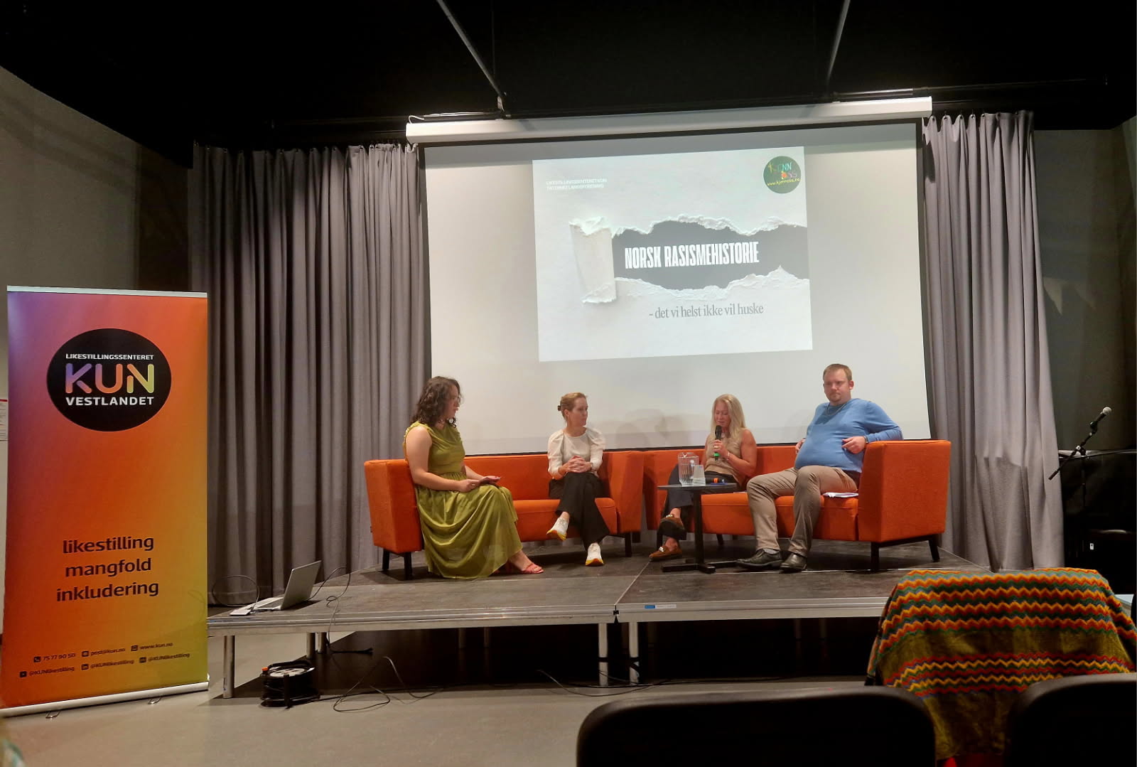 Bildet viser en panelsamtale på en scene med fire personer som sitter i oransje stoler. I bakgrunnen er det en skjerm med teksten "Norsk rasismehistorie - det vi helst ikke vil huske". «KI-generert bildetekst, kvalitetssikret før publisering.»