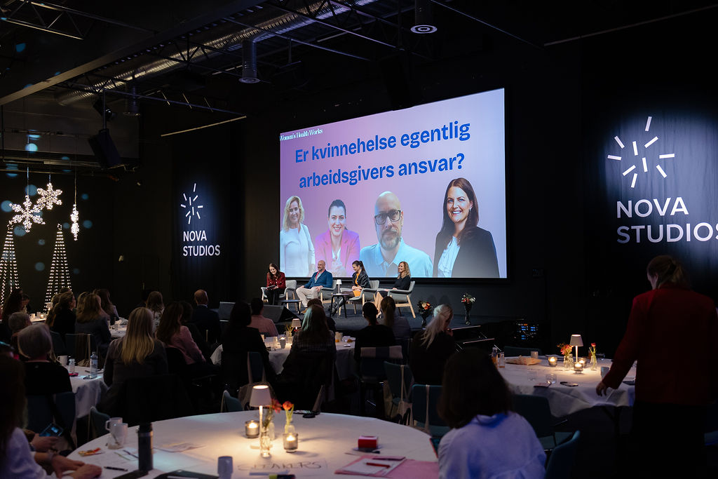 Bildet viser en paneldebatt på Nova Studios med en stor skjerm som har teksten "Er kvinnehelse egentlig arbeidsgivers ansvar?". Flere personer sitter på scenen, mens publikum følger med fra runde bord i salen. Området er dekorert med lys. «KI-generert bildetekst, kvalitetssikret før publisering.»