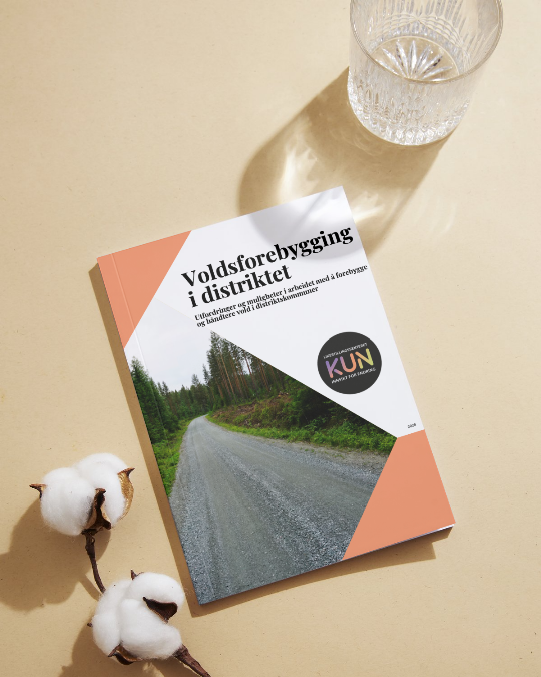 Bildet viser et hefte med tittelen "Voldsforebygging i distriktet" fra Likestillingssenteret KUN. Brosjyren ligger på et bord ved siden av en glassvann og to bomullsdotter. Forsiden av brosjyren har et bilde av en grusvei omgitt av skog. «KI-generert bildetekst, kvalitetssikret før publisering.»