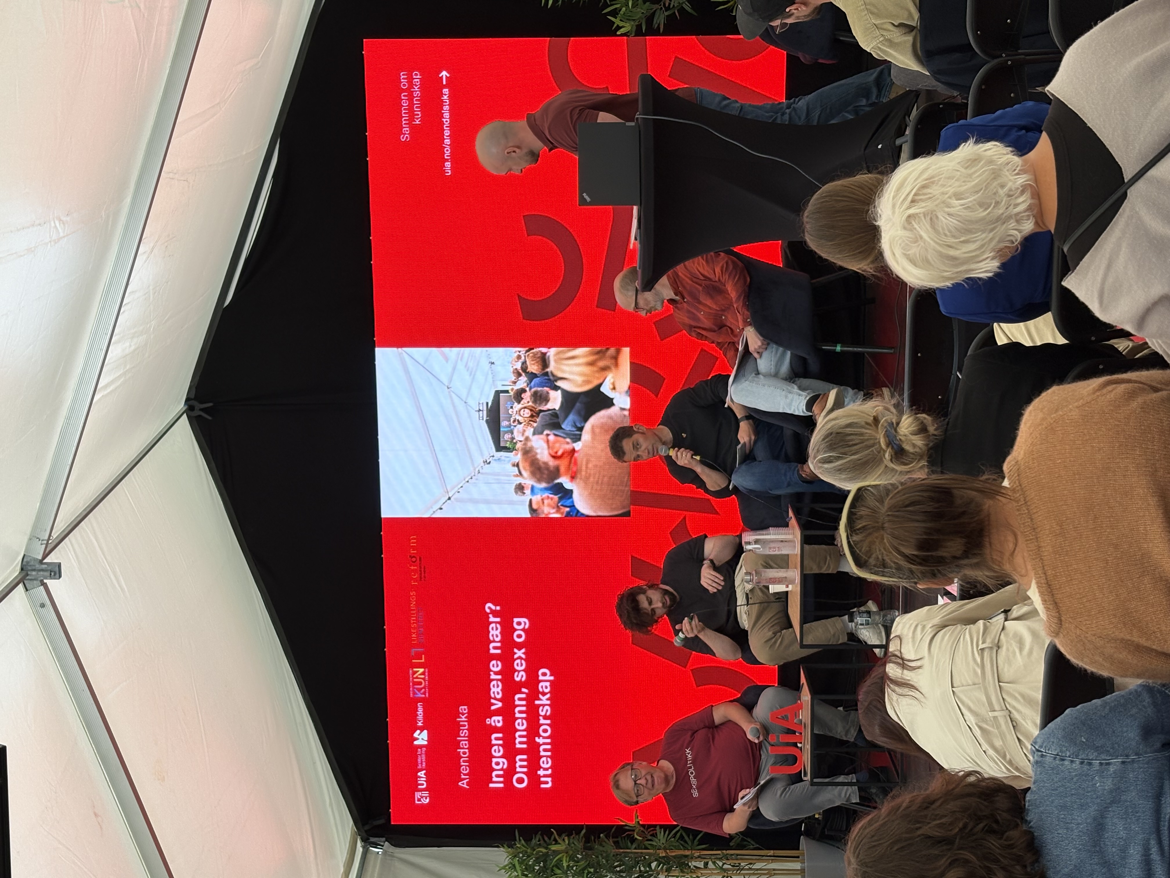 Bildet viser en paneldiskusjon under Arendalsuka med fem deltakere på en scene. Bak dem er en stor skjerm med teksten "Ingen å være nær? Om menn, sex og utenforskap." Publikum sitter foran og hører på. «KI-generert bildetekst, kvalitetssikret før publisering.»