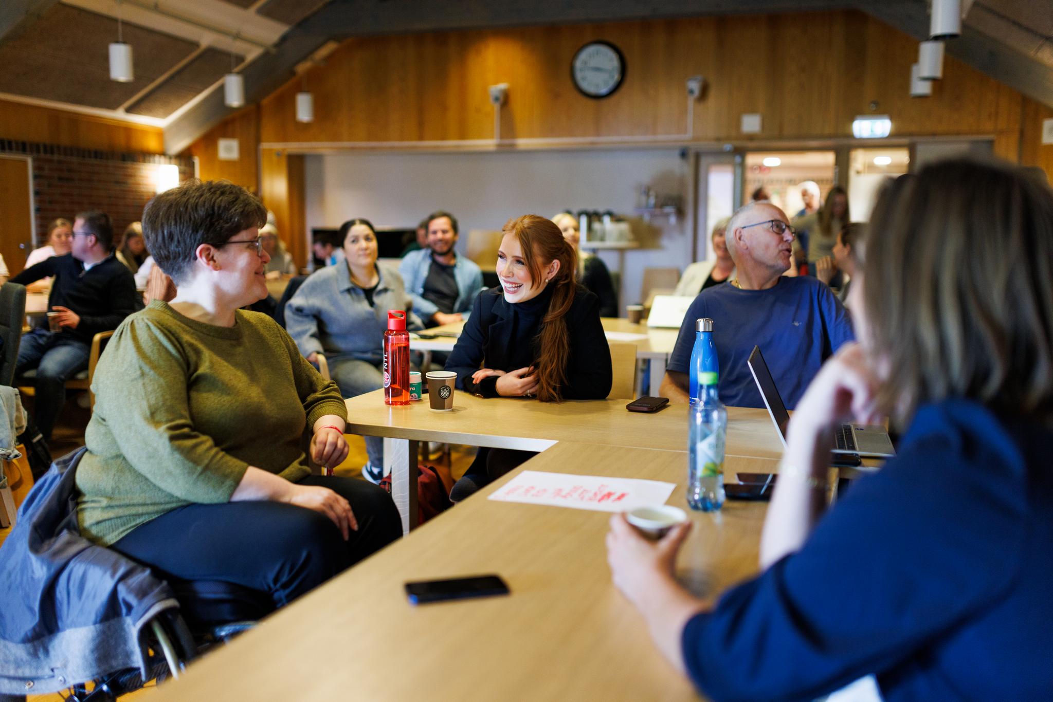 Dette bildet viser en gruppe mennesker som sitter rundt flere bord i et møterom. De ser ut til å være engasjert i en samtale eller en diskusjon og virker avslappede og smilende. Rommet er innredet med trepaneler, og det henger en klokke på veggen. «KI-generert bildetekst, kvalitetssikret før publisering.»