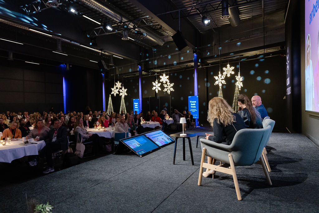 Bildet viser en scene med tre personer som deltar i en paneldiskusjon foran et publikum. Publikum sitter ved runde bord i en stor sal. Det er dekorasjoner med lysende snøfnugg og en skjerm i bakgrunnen. «KI-generert bildetekst, kvalitetssikret før publisering.»