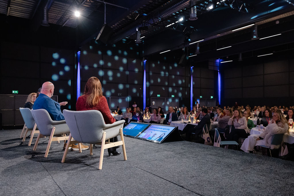 Bildet viser en paneldiskusjon på en scene foran et publikum. Flere personer sitter i stoler på scenen, mens publikum sitter ved runde bord og ser på. Rommet er mørkt med spredte lys og skjermen viser diskusjonsemner. «KI-generert bildetekst, kvalitetssikret før publisering.»