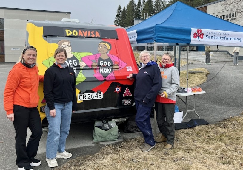 Bildet viser fire personer som står sammen ved siden av en fargerik trailer eller vogn med teksten "DOAVISA" og illustrasjoner med tekstene "JEG ER" og "GOD NOK!". Ved siden av er det et telt med logoen til Norske Kvinners Sanitetsforening, og en utstilt stand ved siden av. Det ser ut som en utendørs arrangement. «KI-generert bildetekst, kvalitetssikret før publisering.»