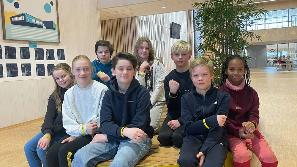 Bildet viser en gruppe barn som sitter sammen innendørs i et lyst rom. De fleste av dem har på seg armbånd, og de ser ut til å ha det hyggelig sammen. Bakgrunnen viser noen kunstverk eller bilder på veggen, og det er planter i nærheten. «KI-generert bildetekst, kvalitetssikret før publisering.»