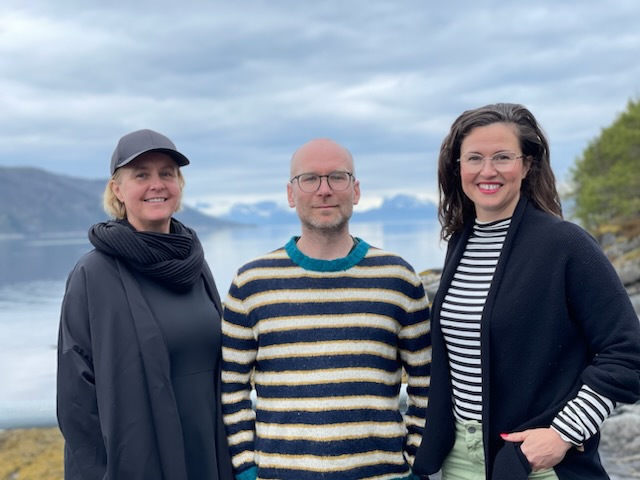 Bildet viser tre personer som står utendørs foran en fjord med fjell i bakgrunnen. De smiler og er kledd i uformelle klær. Det er en overskyet dag, og omgivelsene ser naturskjønne ut. «KI-generert bildetekst, kvalitetssikret før publisering.»