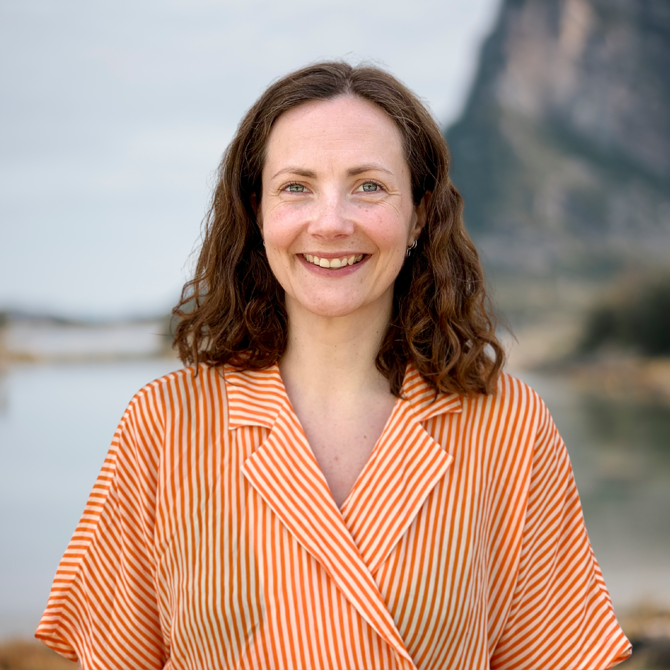 Bildet viser en smilende person med krøllete hår, iført en oransje- og hvitstripete skjorte. Bakgrunnen består av et vassdrag og en fjellformasjon, som gir et naturlig og rolig inntrykk. «KI-generert bildetekst, kvalitetssikret før publisering.»