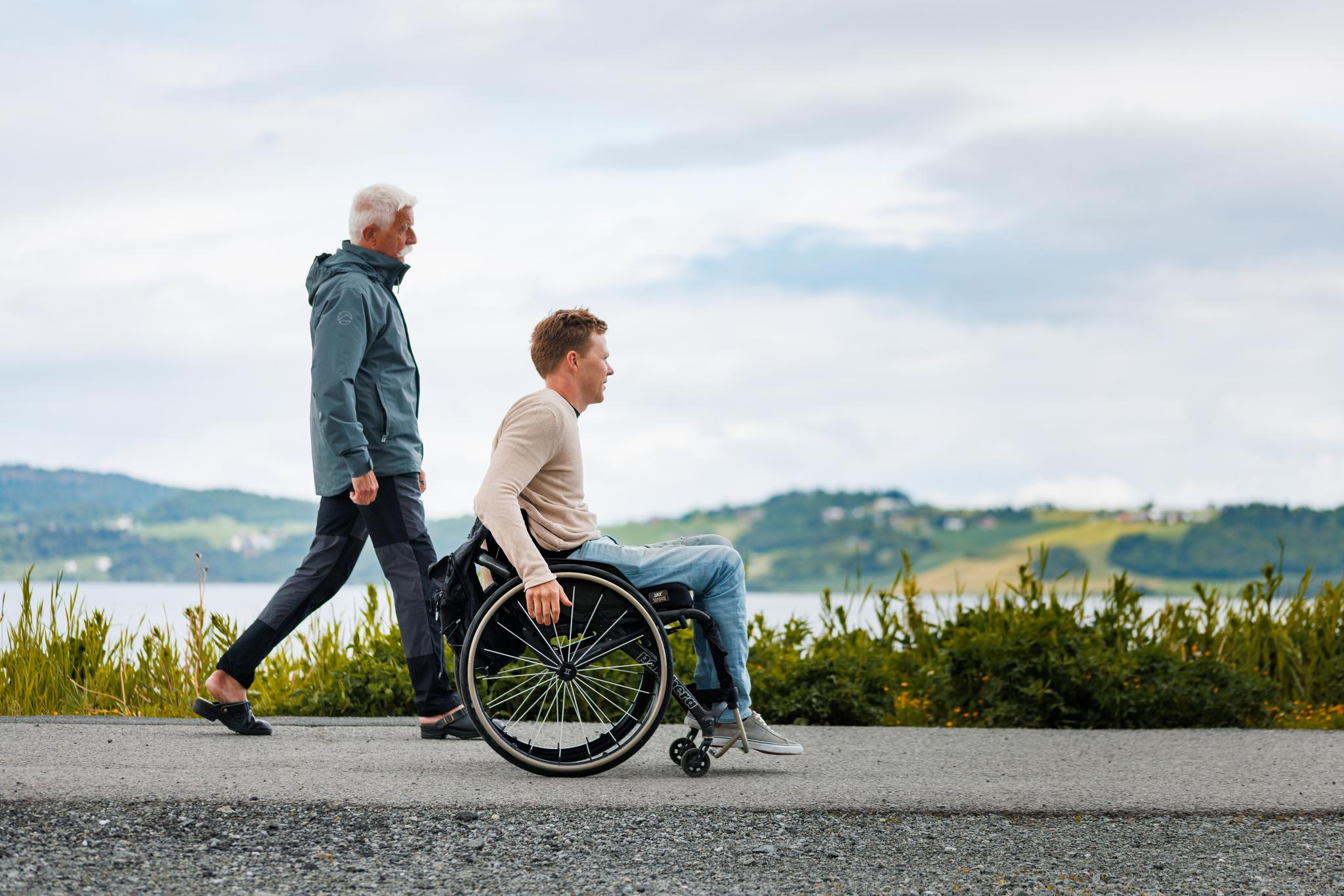 Bildet viser en person i rullestol som er ute på en asfaltert sti ved vannet sammen med en eldre person som går ved siden av. De er i et naturskjønt område med bakgrunn av grønne åser og bebyggelse. «KI-generert bildetekst, kvalitetssikret før publisering.»