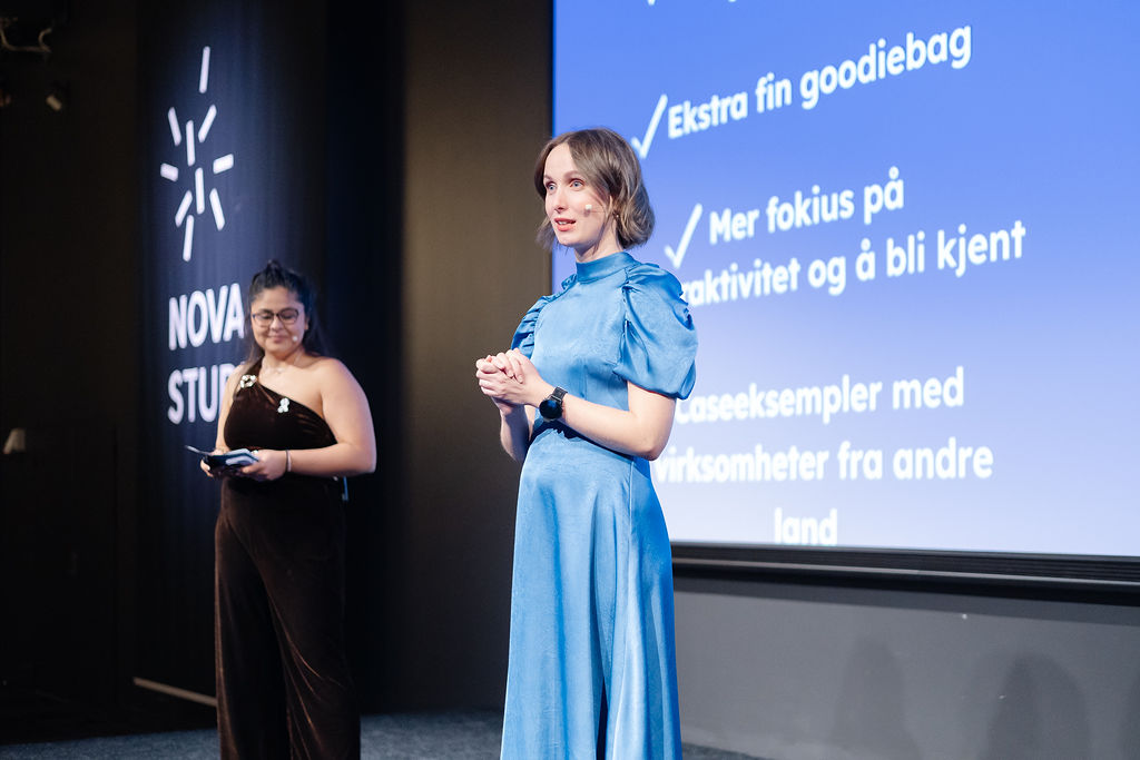 To personer står på en scene foran en skjerm med tekst. Bak dem vises punkter om aktiviteter og eksempler fra andre land. «KI-generert bildetekst, kvalitetssikret før publisering.»