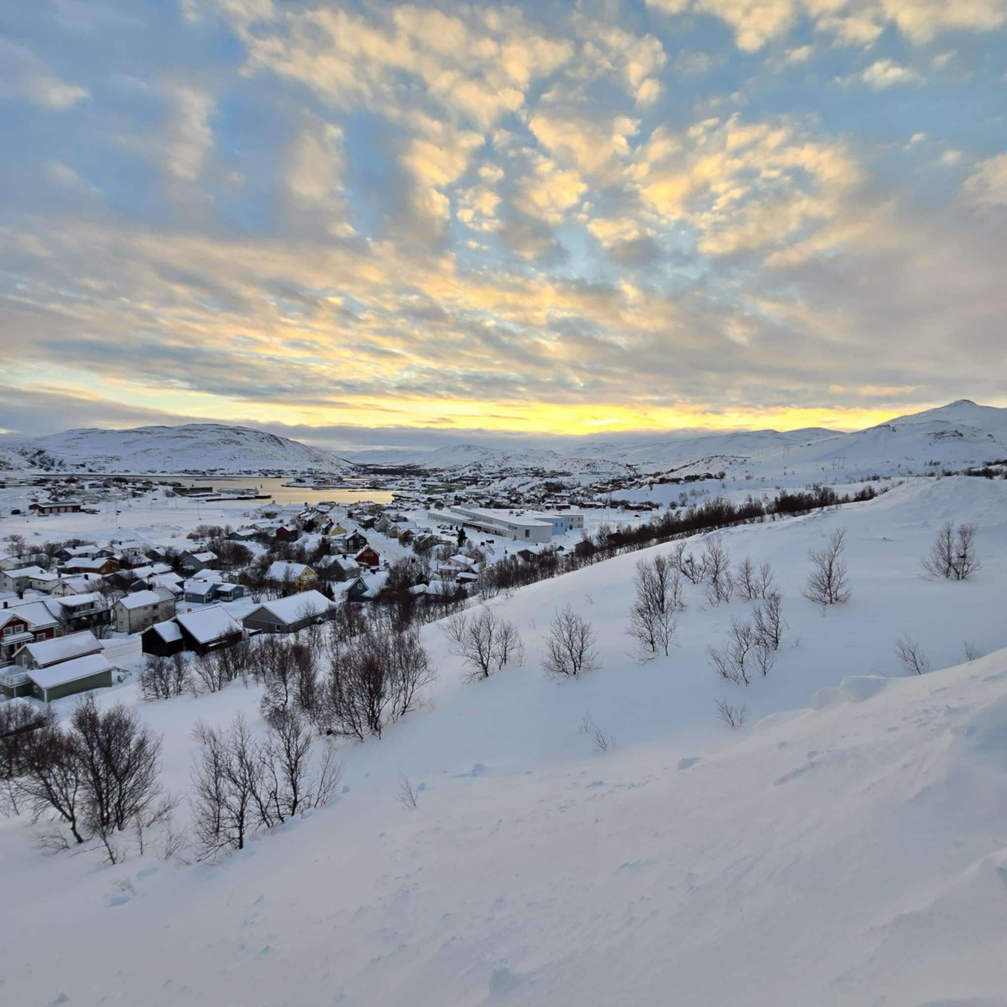 Bildet viser et snødekt landskap med en liten by eller tettsted i dalen. Husene er omgitt av snø, og noen trær stikker opp av snøen. I bakgrunnen ses fjell, og himmelen er delvis skyet med gylne farger fra solnedgangen eller soloppgangen. «KI-generert bildetekst, kvalitetssikret før publisering.»