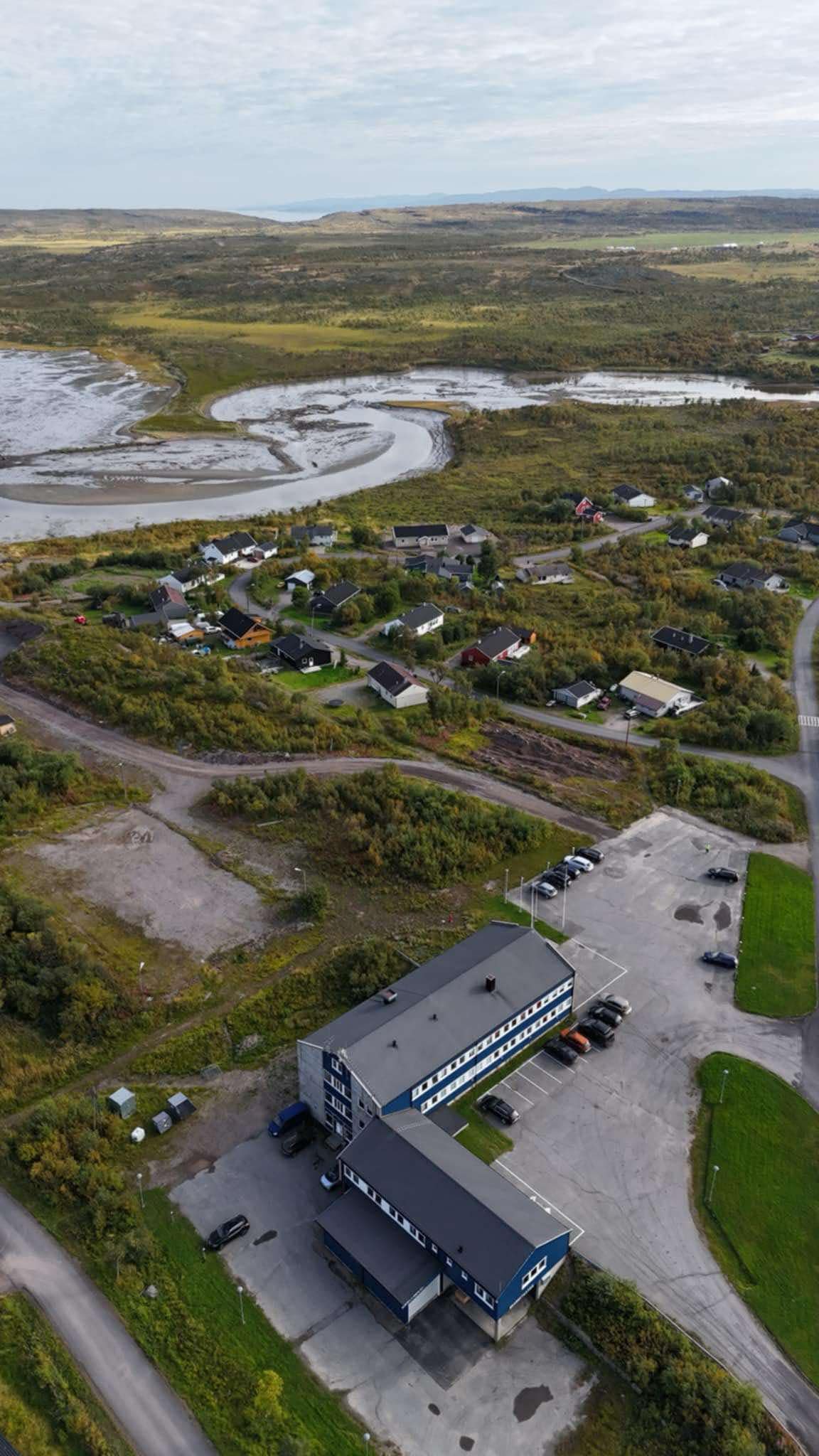 Bildet viser et lite tettsted med spredt bebyggelse omgitt av natur. Forgrunnen domineres av en større bygning omgitt av parkeringsplass. I bakgrunnen kan man se elver og grønt landskap under en noe skyet himmel. «KI-generert bildetekst, kvalitetssikret før publisering.»