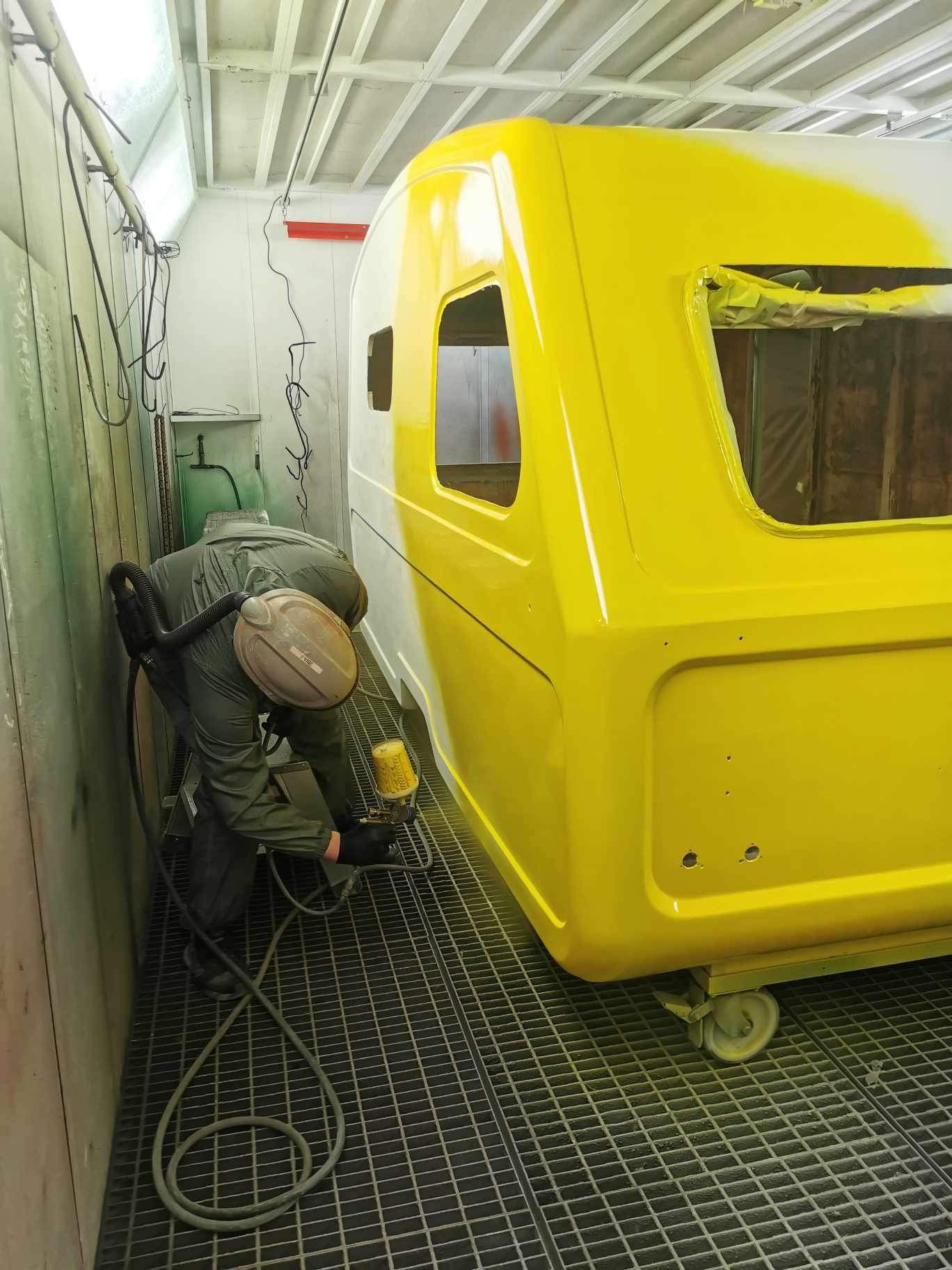 En person sprøytelakkerer en lys gul campingvogn i et verksted. Personen har på seg beskyttelsesutstyr, inkludert hansker og maske. Verkstedet har rutenettgulv og ventilasjon i taket.  «KI-generert bildetekst, kvalitetssikret før publisering.»