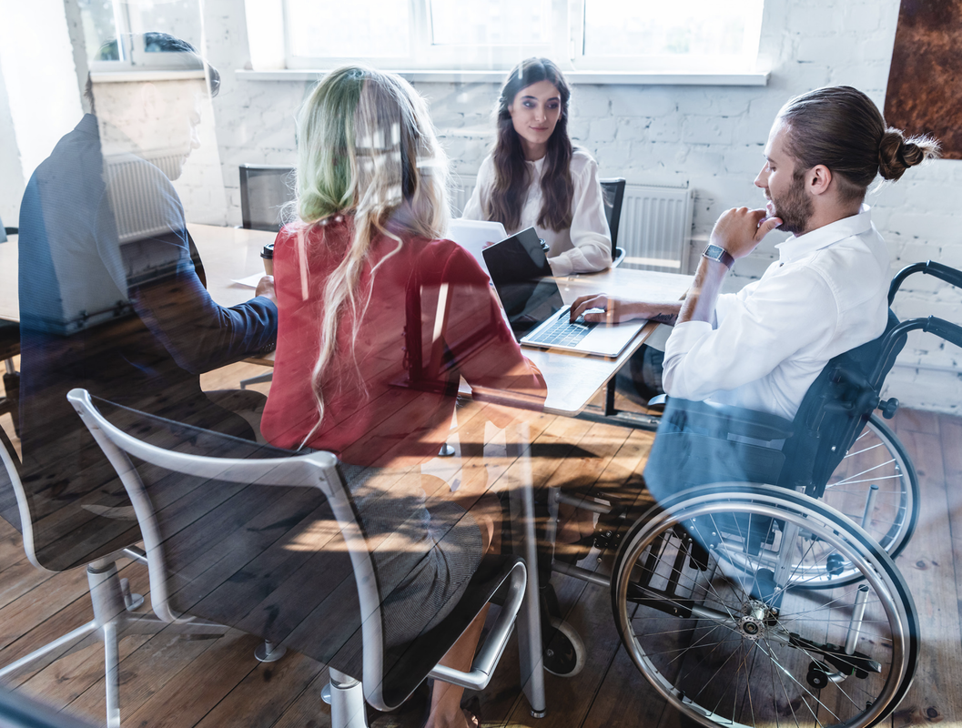 Bildet viser en gruppe mennesker som sitter rundt et konferansebord. De ser ut til å ha et møte eller en diskusjon, og én person sitter i rullestol. Refleksjoner i vinduet gir en dynamisk effekt.