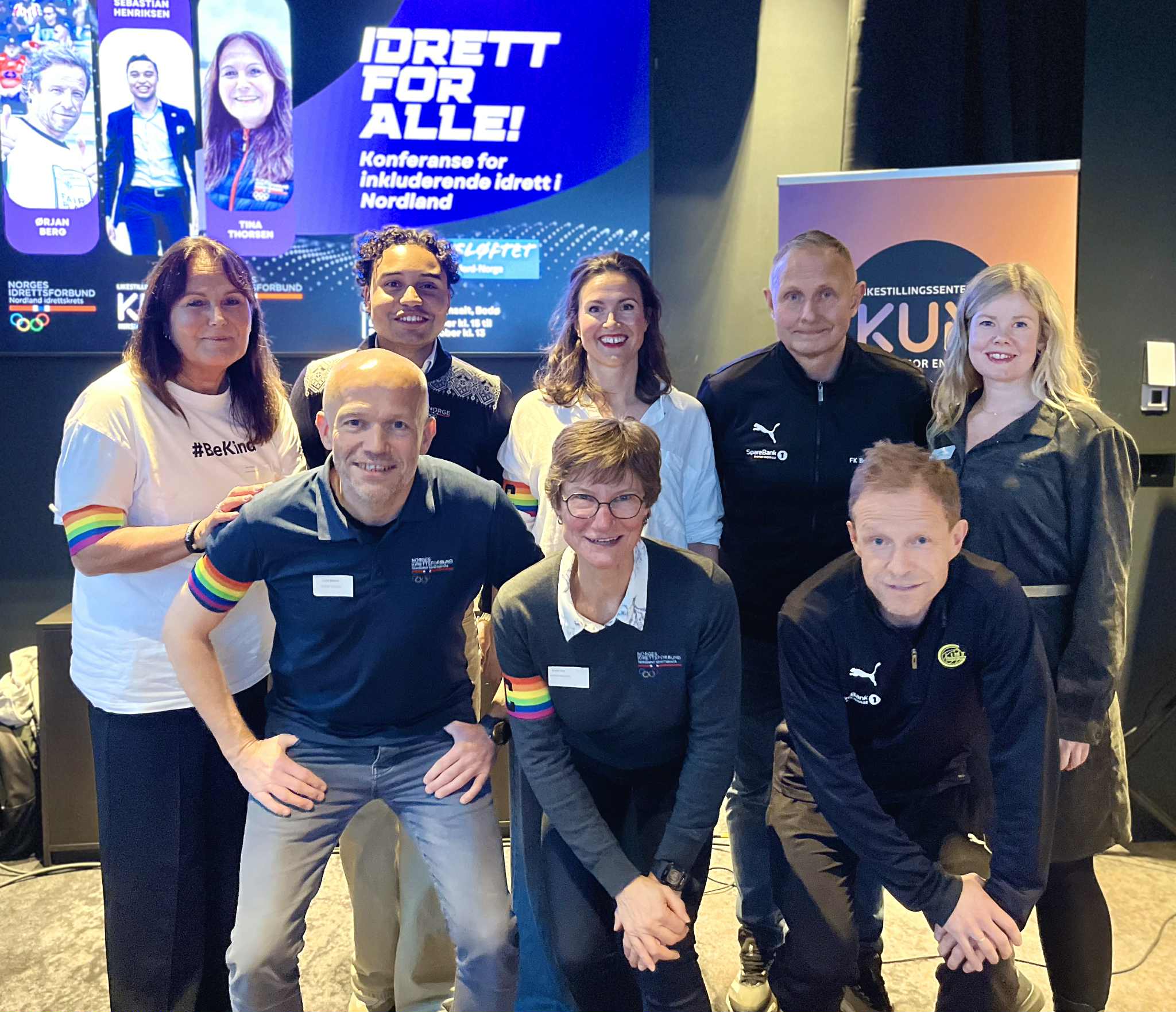 Dette bildet viser en gruppe mennesker som er samlet foran en plakat med teksten "Idrett for alle! Konferanse for inkluderende idrett i Nordland". De fleste har på seg klær med regnbuefarger som symbol på inkludering og mangfold. «KI-generert bildetekst, kvalitetssikret før publisering.»