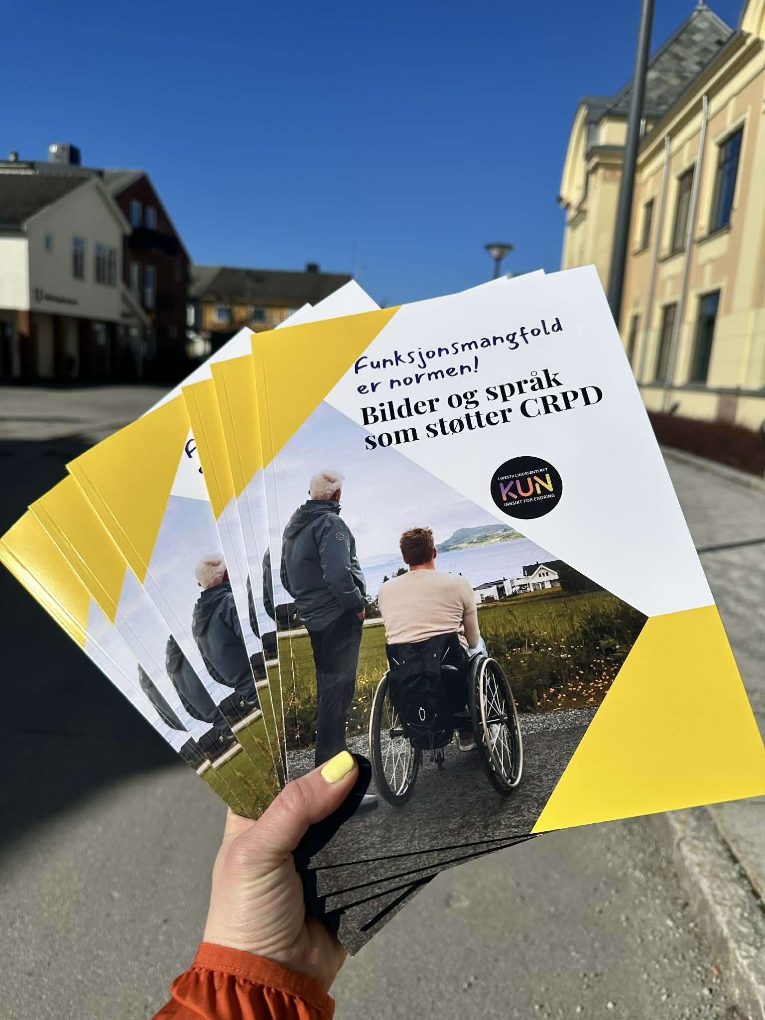 Bildet viser en hånd som holder en bunke brosjyrer. På forsiden av brosjyrene står det "Funksjonsmangfold er normen! Bilder og språk som støtter CRPD." Brosjyrene har et bilde av to personer, hvorav en er i rullestol og en annen står ved siden av. Dette foregår i et utendørs miljø, og det er en klar blå himmel i bakgrunnen. «KI-generert bildetekst, kvalitetssikret før publisering.»