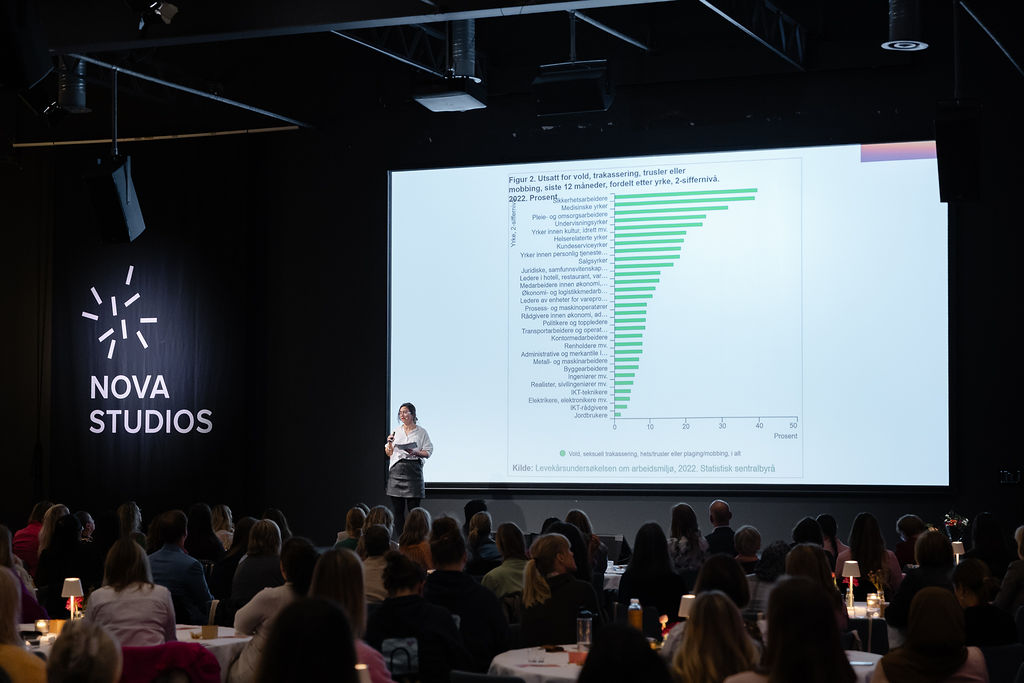 En person holder en presentasjon foran et publikum i et auditorium. Det vises en graf på skjermen, og lokalet er merket med "NOVA STUDIOS". Publikum sitter ved bord med stearinlys. «KI-generert bildetekst, kvalitetssikret før publisering.»