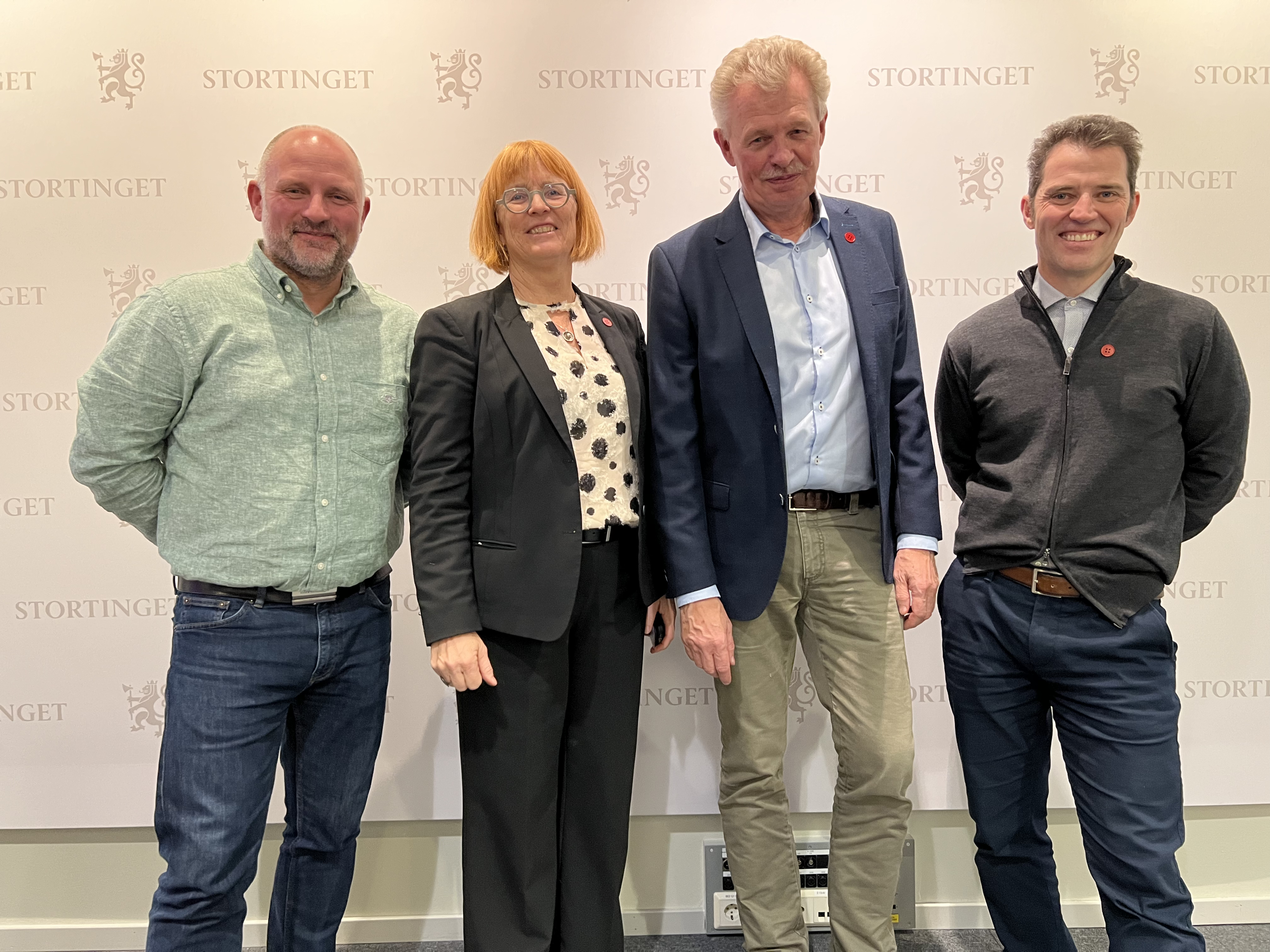Bildet viser fire personer som står foran en bakgrunn med Stortingets logo. De ser ut til å være formelt kledd og poserer for et gruppebilde. «KI-generert bildetekst, kvalitetssikret før publisering.» 