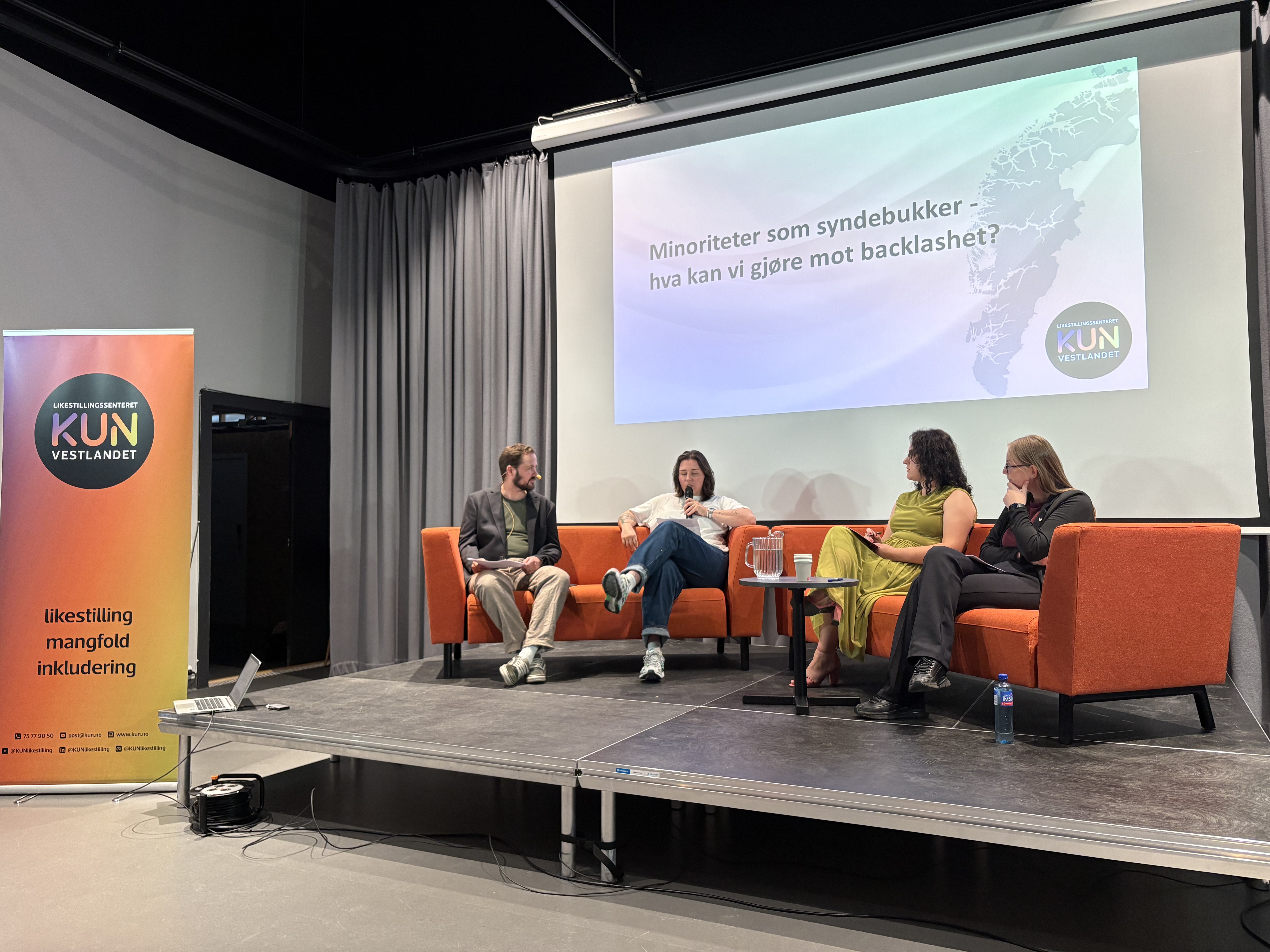 Bildet viser et panel med fire personer som sitter i oransje stoler på en scene. En person snakker i en mikrofon. Bak dem er en skjerm hvor det står "Minoriteter som syndebukker – hva kan vi gjøre mot backlashen?". Til venstre er en rollup for Likestillingssenteret på Vestlandet.