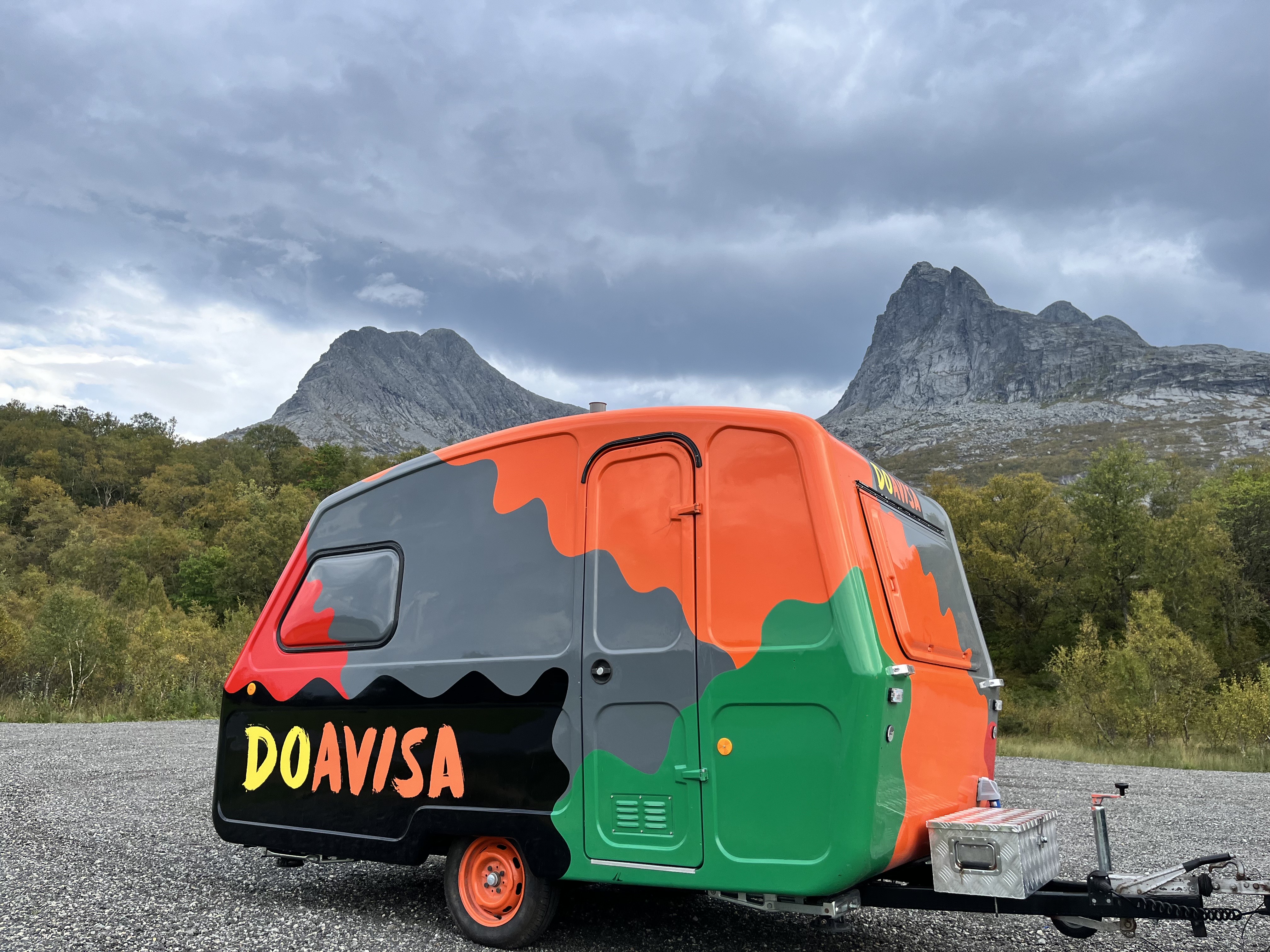 Bildet viser en fargerik campingvogn med teksten "DOAVISA" på siden, foran et fjellandskap med skyer.  «KI-generert bildetekst, kvalitetssikret før publisering.»