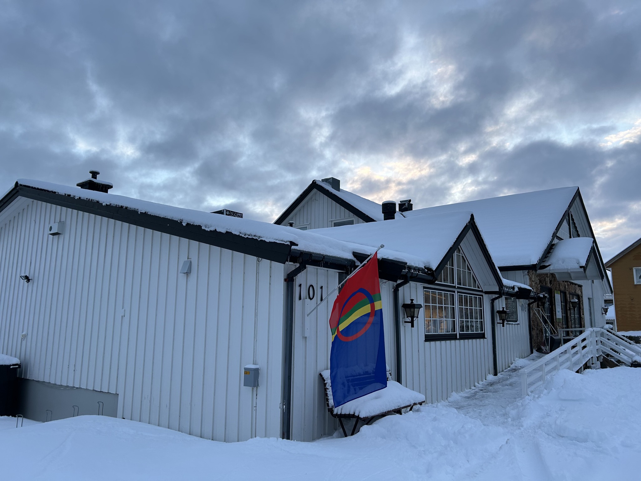 Bildet viser et hvitt bygg dekket av snø, med et samisk flagg som vaier foran. Himmelen er overskyet, og det ligger snø på bakken og takene.