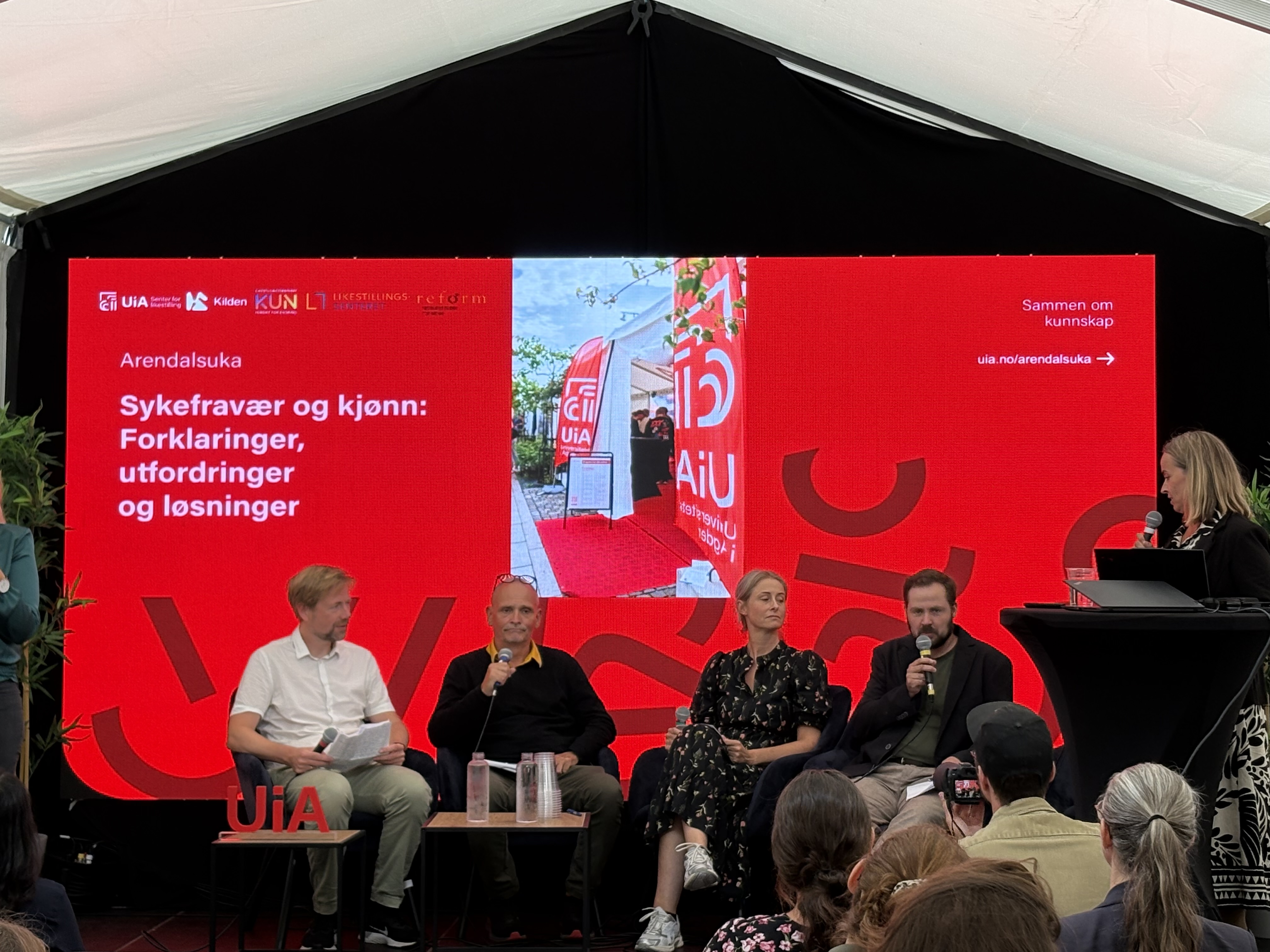 Bildet viser en paneldiskusjon under Arendalsuka. Temaet er "Sykefravær og kjønn: Forklaringer, utfordringer og løsninger". Det er en stor rød bakgrunn med tekst, og flere personer sitter eller står ved et podium og deltar i samtalen. Publikum ser på. «KI-generert bildetekst, kvalitetssikret før publisering.»