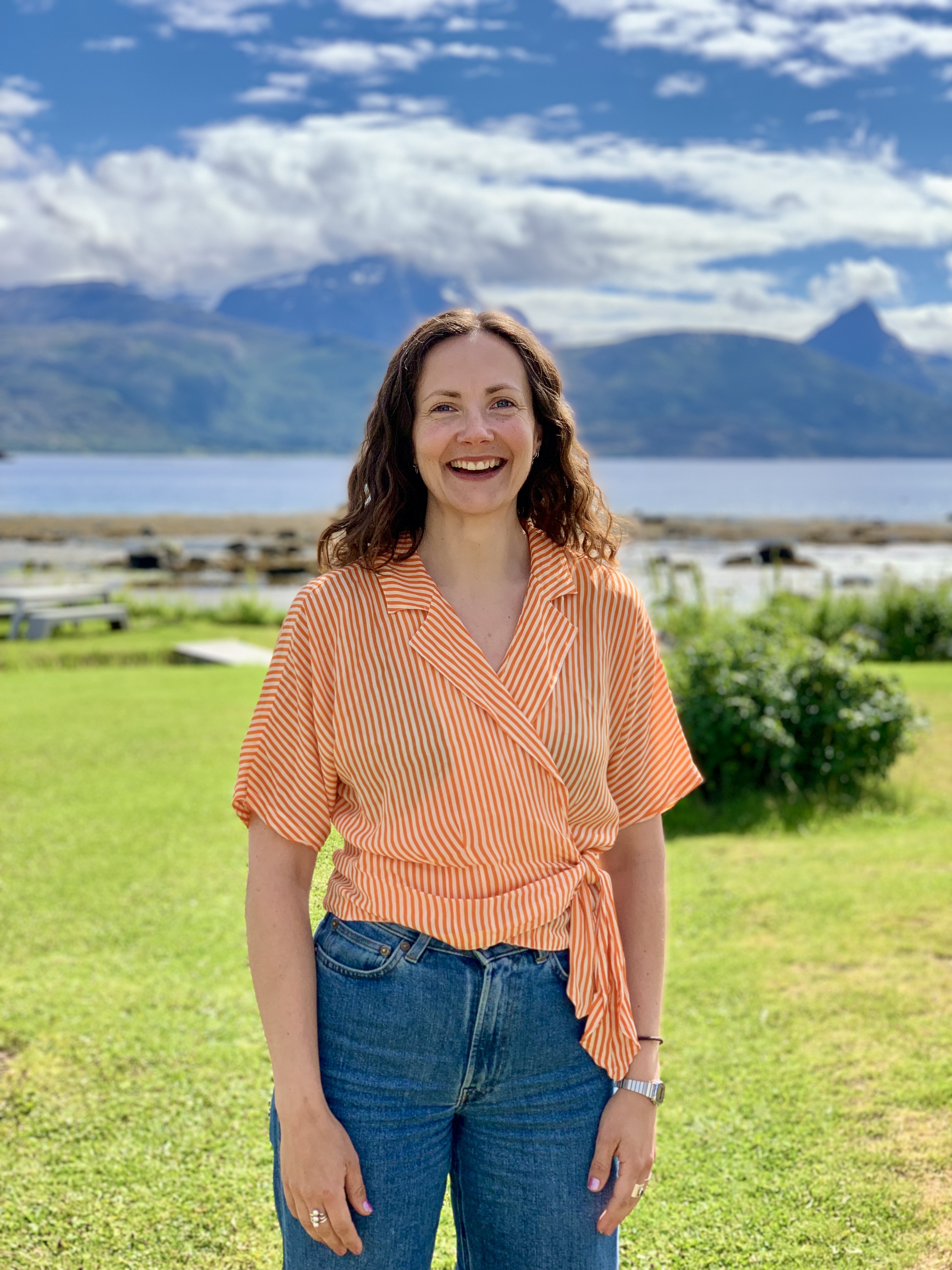 Bildet viser en person som smiler, stående utendørs med en vakker naturbakgrunn bestående av fjell, vann og grønt gress. Personen har på seg en stripete, oransje topp og jeans. Været ser fint ut med blå himmel og noen skyer. «KI-generert bildetekst, kvalitetssikret før publisering.»