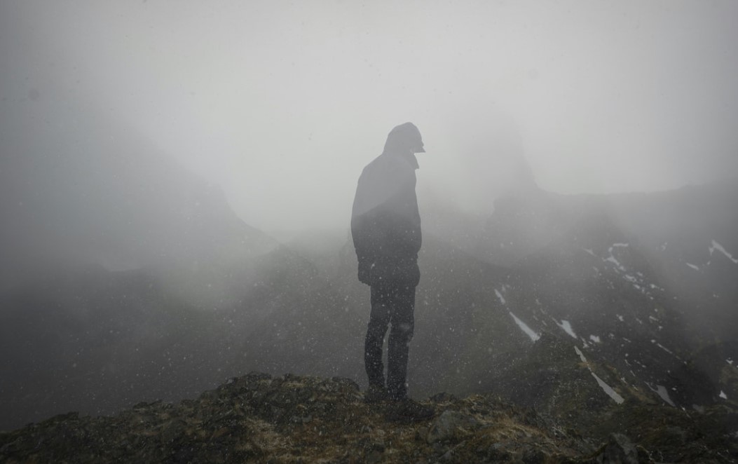 Mann som står på fjellet i dårlig vær. Han står med ryggen til kameraet. 