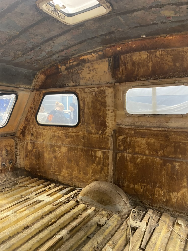 Bildet viser interiøret i et gammelt kjøretøy, trolig en varebil eller bobil, med avskallet maling og slitt metal. Vinduer med utsikt til personer utenfor er synlige, og gulvet ser ut til å være delvis rustent. «KI-generert bildetekst, kvalitetssikret før publisering.»