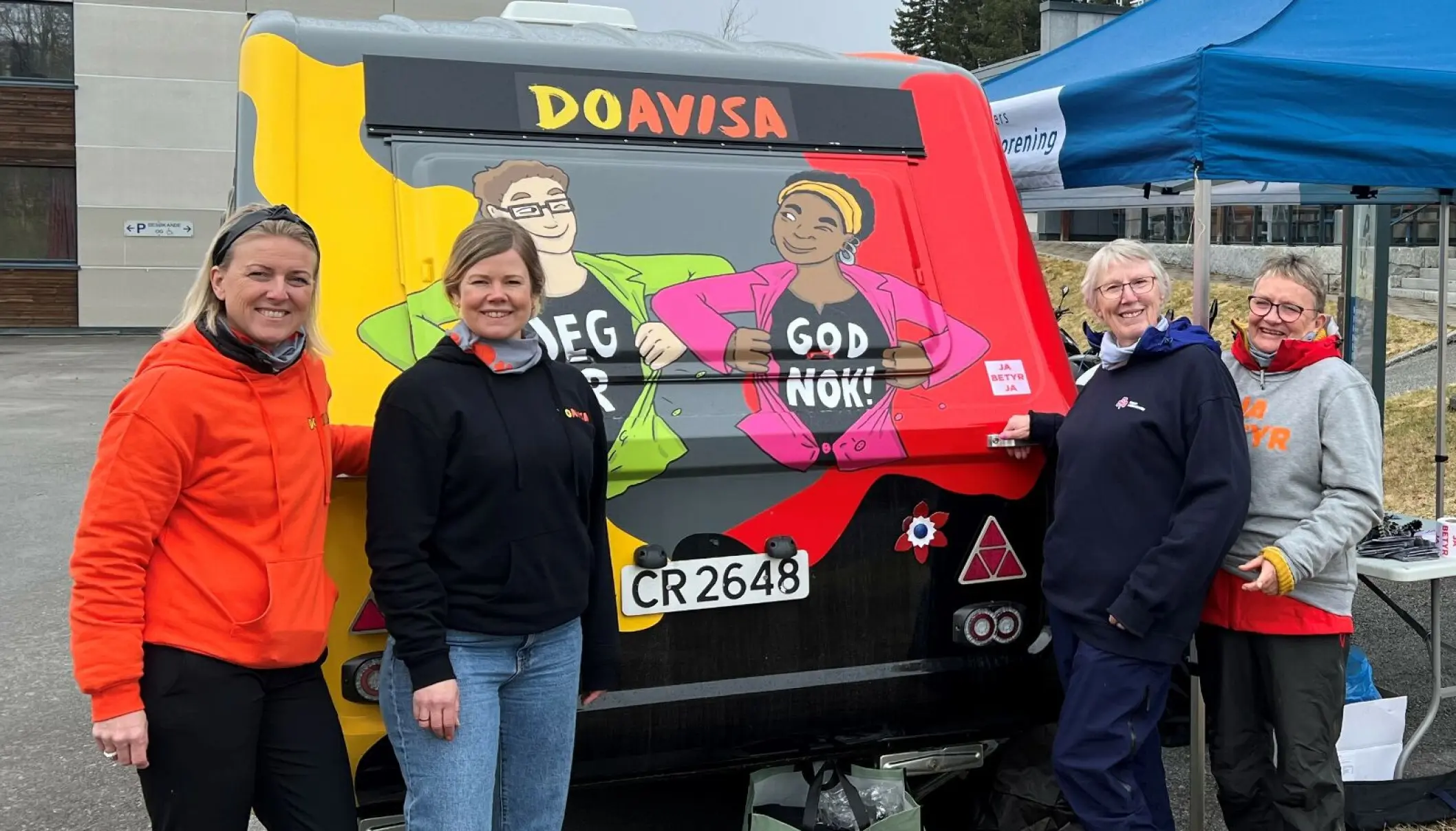 Fire personer står ved siden av en fargerik campingvogn med teksten "DOAVISA" og illustrasjoner med budskapene "JEG ER" og "GOD NOK!" på. De smiler, og det ser ut til å være en uformell og positiv situasjon, kanskje i forbindelse med et arrangement eller en kampanje. «KI-generert bildetekst, kvalitetssikret før publisering.» 