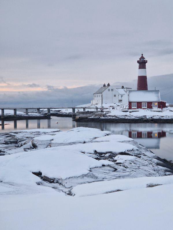 Bildet viser et snødekt landskap med et fyrtårn og hus ved vannet. Det er en bro i bakgrunnen, og himmelen har et svakt lys fra solen.