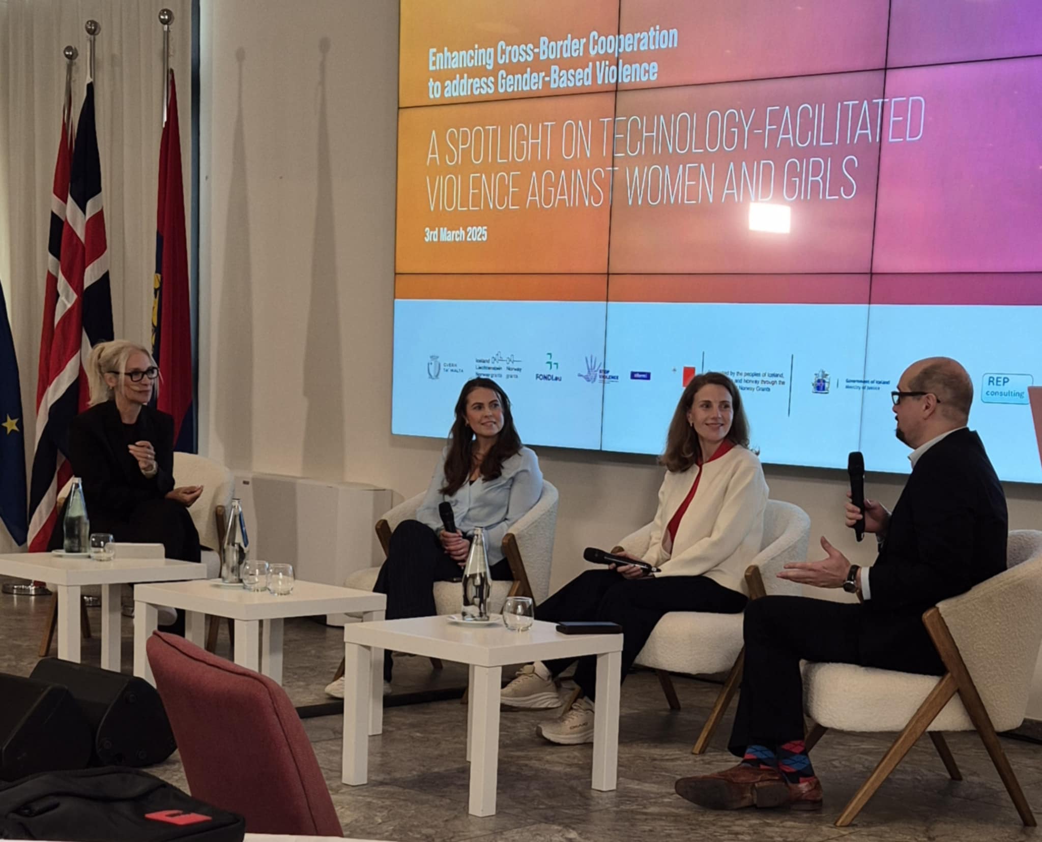 Bildet viser en paneldiskusjon med fire deltakere som sitter i stoler på en scene. Bak dem er det en stor skjerm med teksten “Enhancing Cross-Border Cooperation to address Gender-Based Violence” og “A Spotlight on Technology-Facilitated Violence Against Women and Girls”, datert 3. mars 2025. Det er diverse flagg i bakgrunnen. Deltakerne holder mikrofoner og er engasjert i samtalen. «KI-generert bildetekst, kvalitetssikret før publisering.»