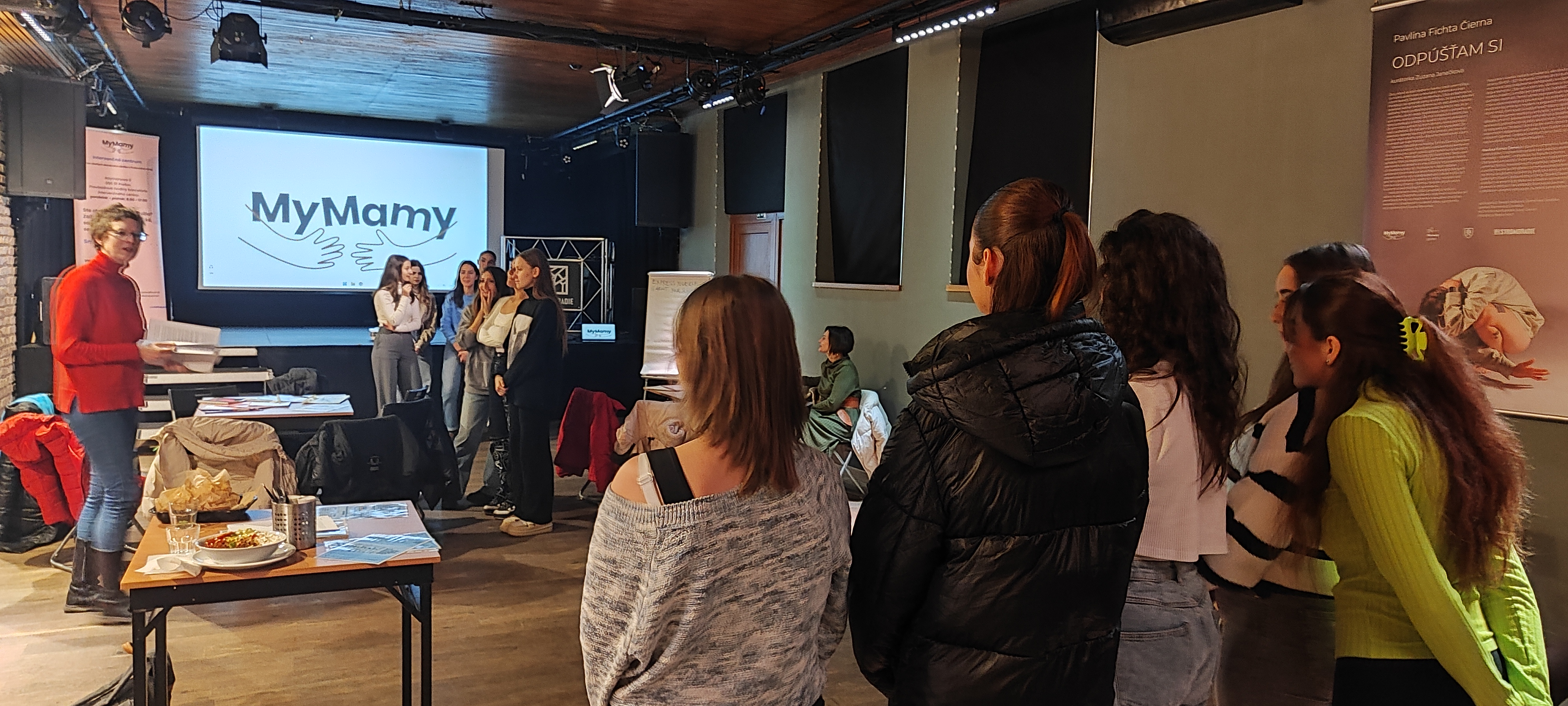 Dette bildet viser en gruppe mennesker samlet i et rom med en presentasjon på en skjerm som viser teksten "MyMamy." Folk ser ut til å delta i en slags workshop eller møte. Rommet har et uformelt oppsett, med stoler, bord med mat og papir, og det er interaksjon mellom deltakerne.