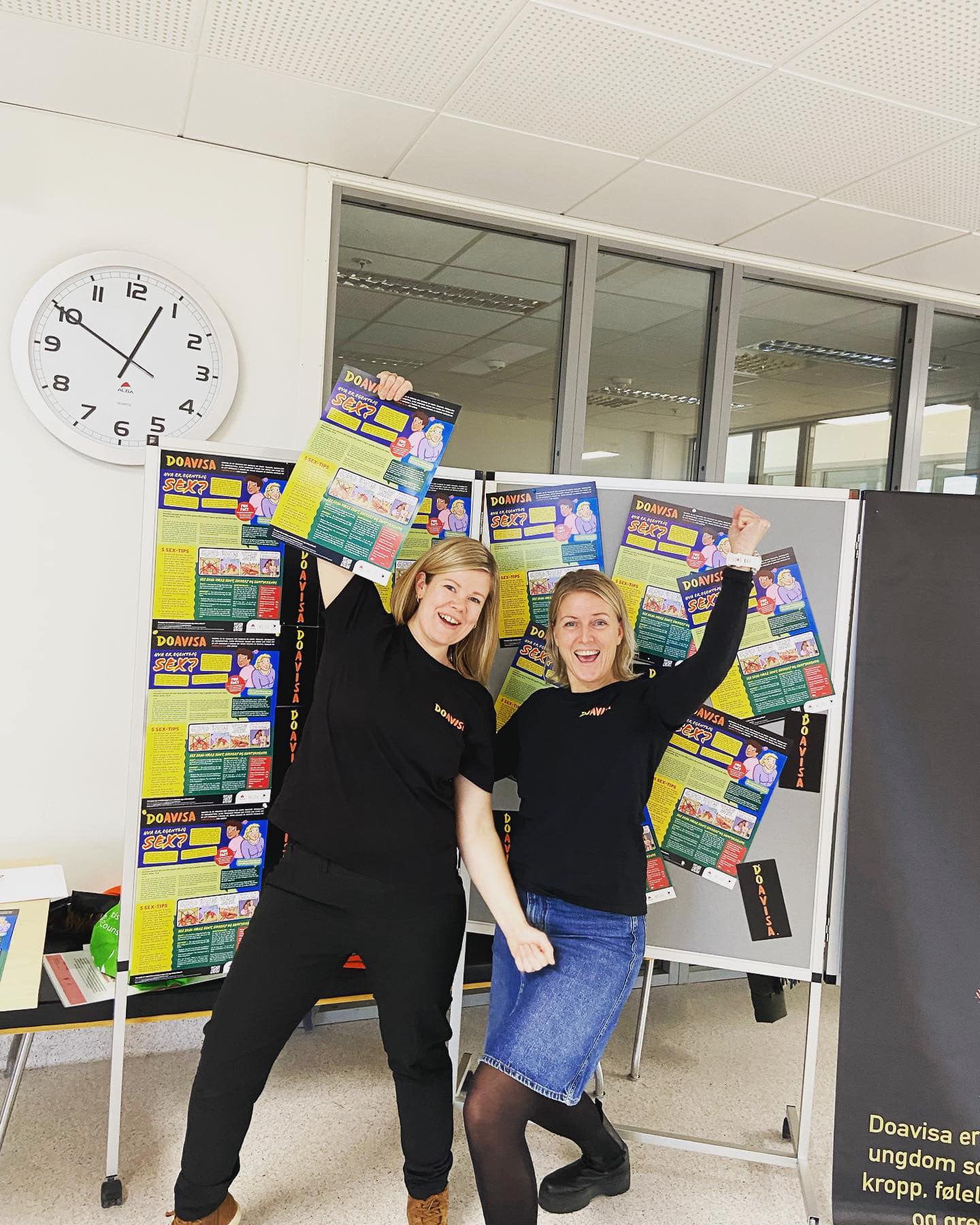 To personer poserer gledelig foran en vegg dekket med fargerike plakater. De holder opp en plakat hver, og det er en klokke på veggen i bakgrunnen. «KI-generert bildetekst, kvalitetssikret før publisering.» 