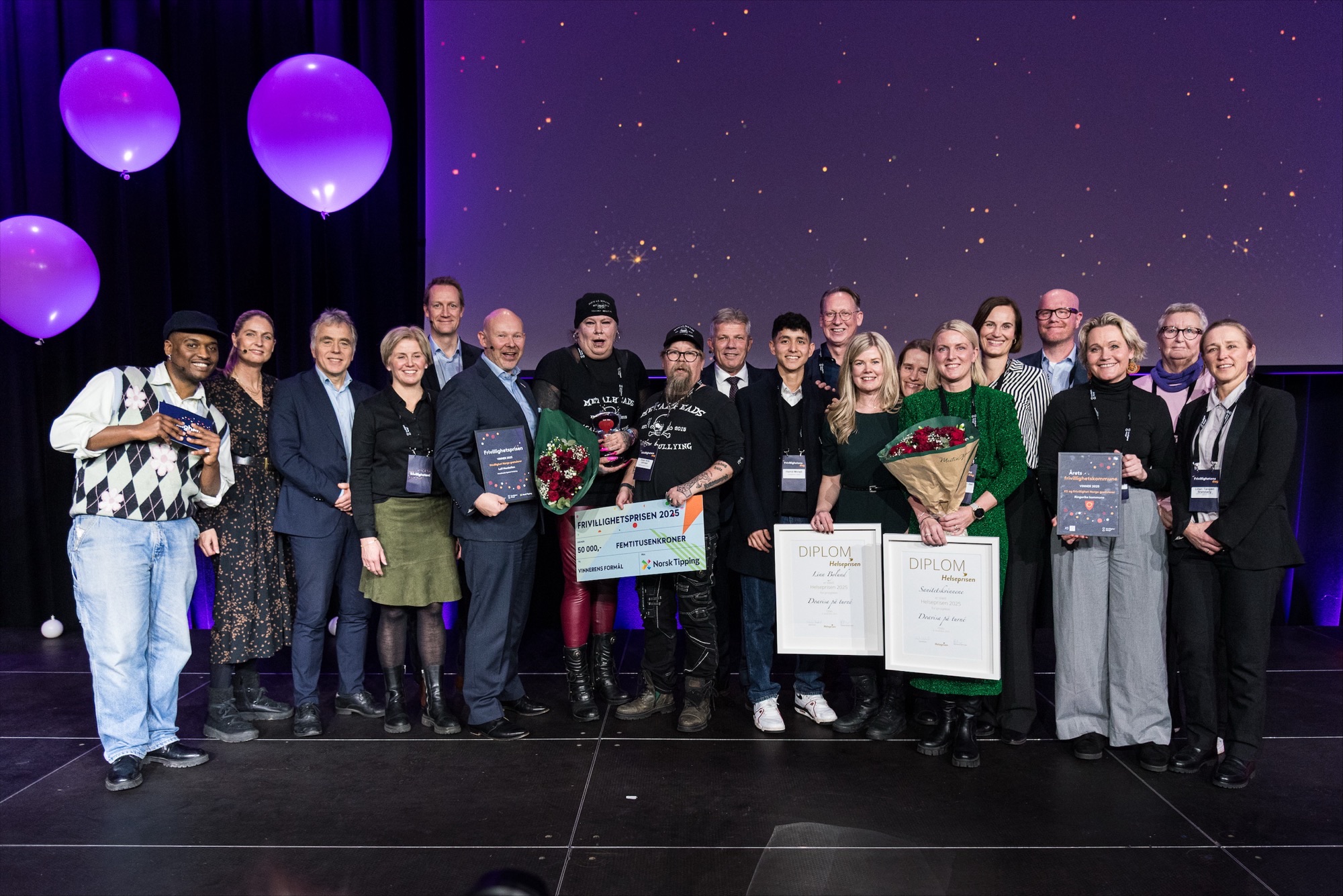 Bildet viser en stor gruppe mennesker som poserer sammen på en scene. Noen holder diplomer og blomsterbuketter. Det er ballonger i bakgrunnen, og stemningen ser ut til å være festlig. Alle de glade vinnerne av de nasjonale frivillighetsprisene, sammen med prisutdelerne på Sentralen i Oslo. 