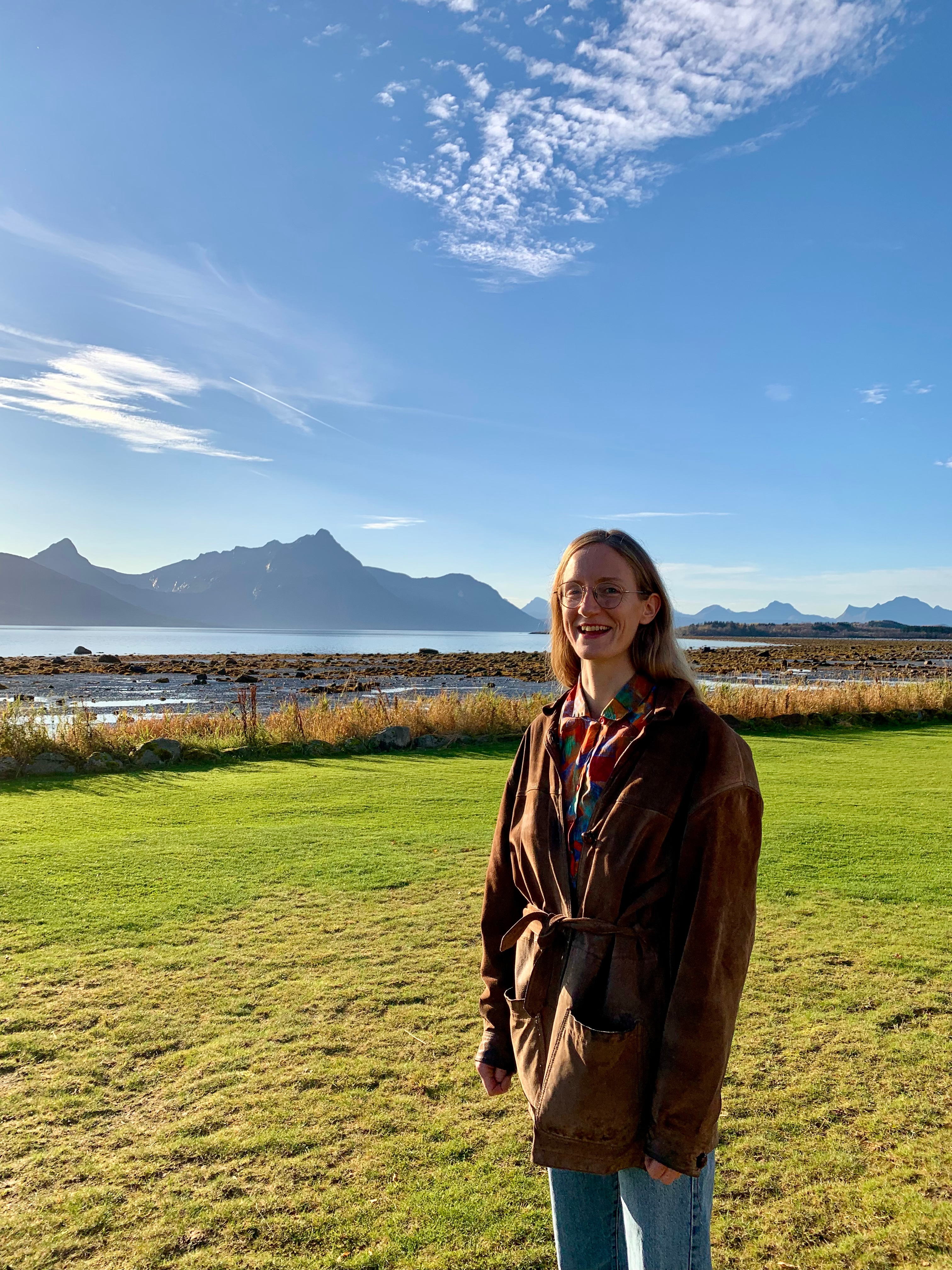 Bildet viser en person som står på en gressplen med en vakker naturbakgrunn. Det er fjell og vann i bakgrunnen under en klar blå himmel med noen skyer. Personen smiler og har på seg en brun jakke og briller.