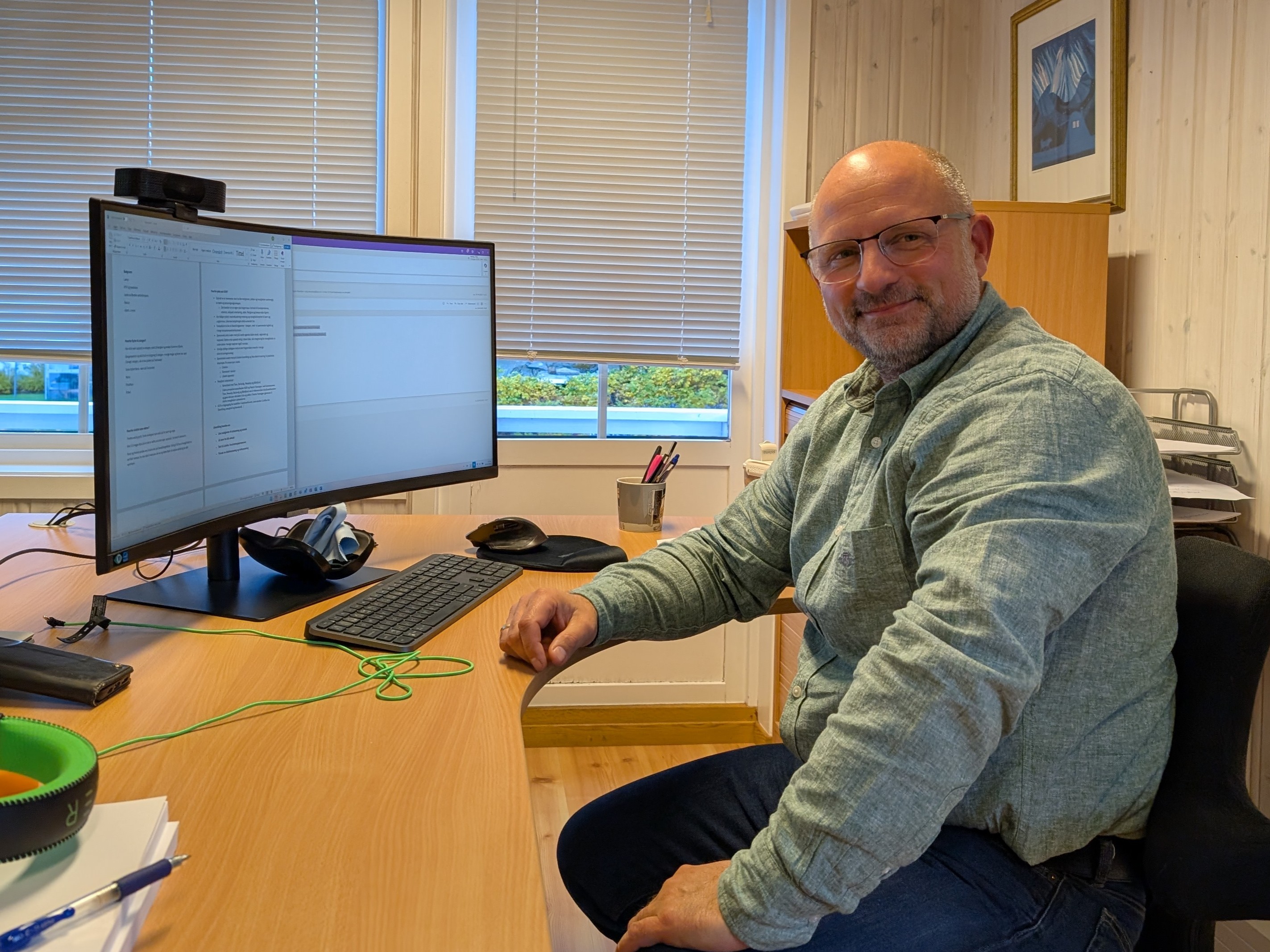 En person sitter ved et skrivebord med en stor skjerm, tastatur og mus foran seg. Personen har på seg en grønn skjorte og ser mot kameraet med et smil. Kontoret har lukkede persienner og en innrammet illustrasjon på veggen. «KI-generert bildetekst, kvalitetssikret før publisering.» 