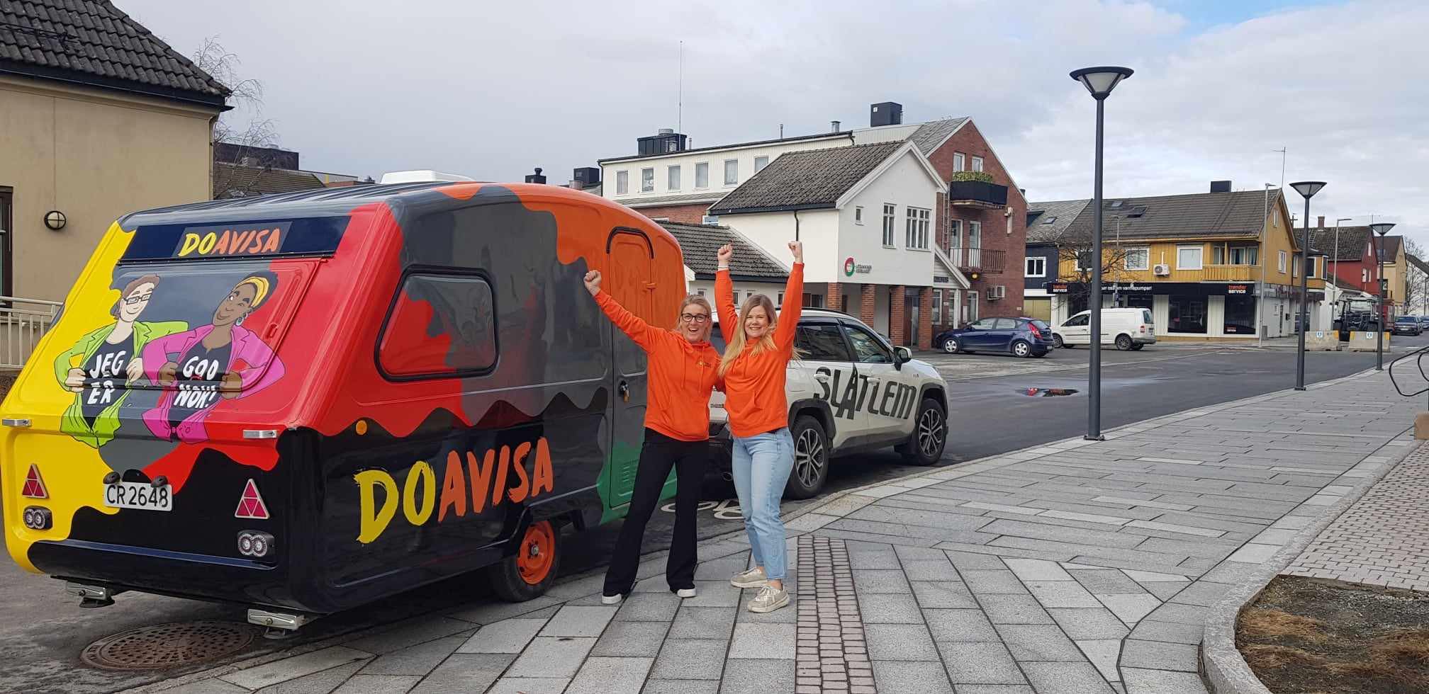 Bildet viser to personer i oransje gensere som står ved siden av en fargerik campingvogn med teksten "DOAVISA." Vognen er festet til en bil, og bakgrunnen viser en urban gate med bygninger. «KI-generert bildetekst, kvalitetssikret før publisering.»