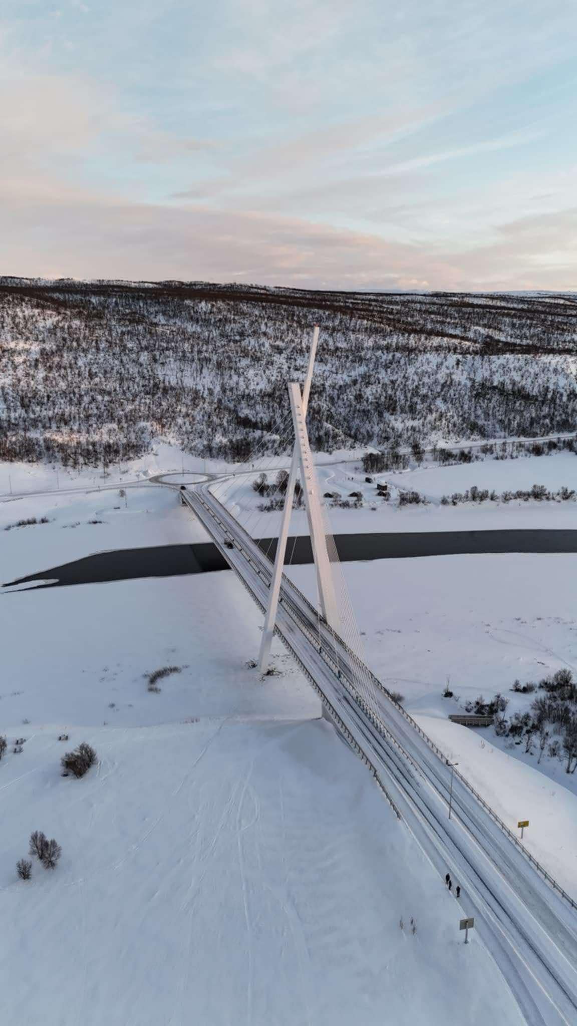 Dette bildet viser en moderne bro som krysser en delvis frossen elv i et snødekket landskap. Broen har en unik design med skråstilte pilarer og stålwire. Området rundt er dekket av snø, og det er noen busker og trær som stikker opp. Himmlen er blå med noen skyer. «KI-generert bildetekst, kvalitetssikret før publisering.»