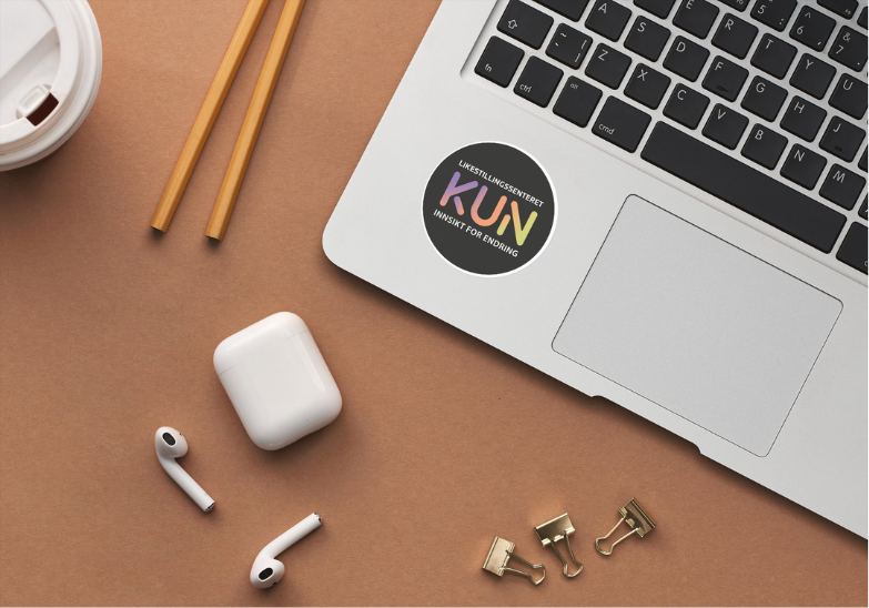 Bildet viser en bærbar datamaskin med et klistremerke som har teksten "Likestillingssenteret KUN med visjon - Innsikt for Endring". Rundt datamaskinen er en kaffekopp, to blyanter, et par trådløse ørepropper, en øreproppetui, og noen papirklips på et brunt underlag. «KI-generert bildetekst, kvalitetssikret før publisering.»