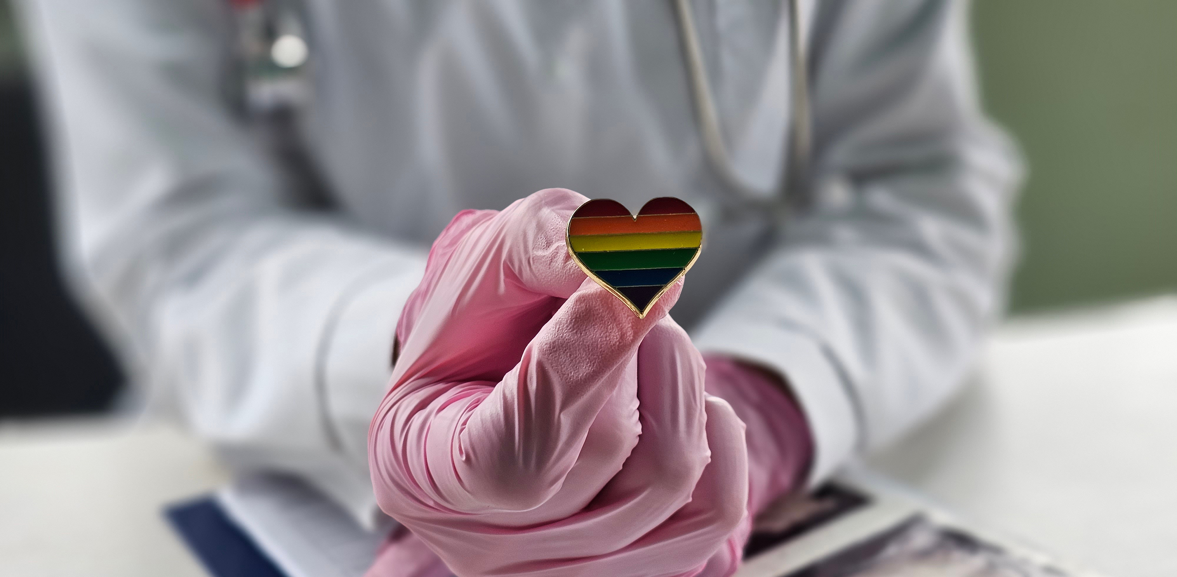 Bildet viser en person med rosa hansker i en hvit frakk som holder en hjerteformet pin med regnbuemønster. «KI-generert bildetekst, kvalitetssikret før publisering.»