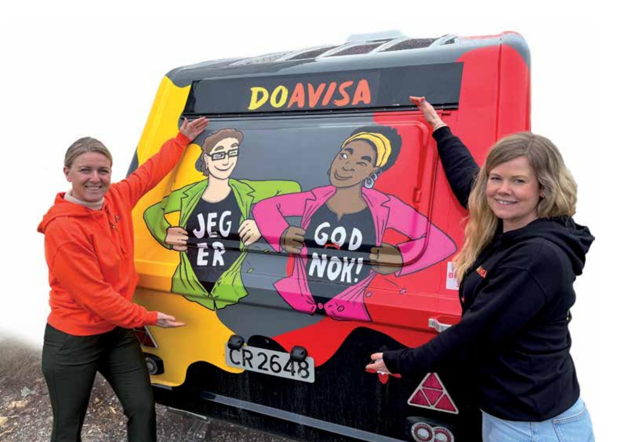 Bildet viser to smilende personer i fargerike klær bak en campingvogn med kunstverk som har teksten "JEG ER GOD NOK!". «KI-generert bildetekst, kvalitetssikret før publisering.»