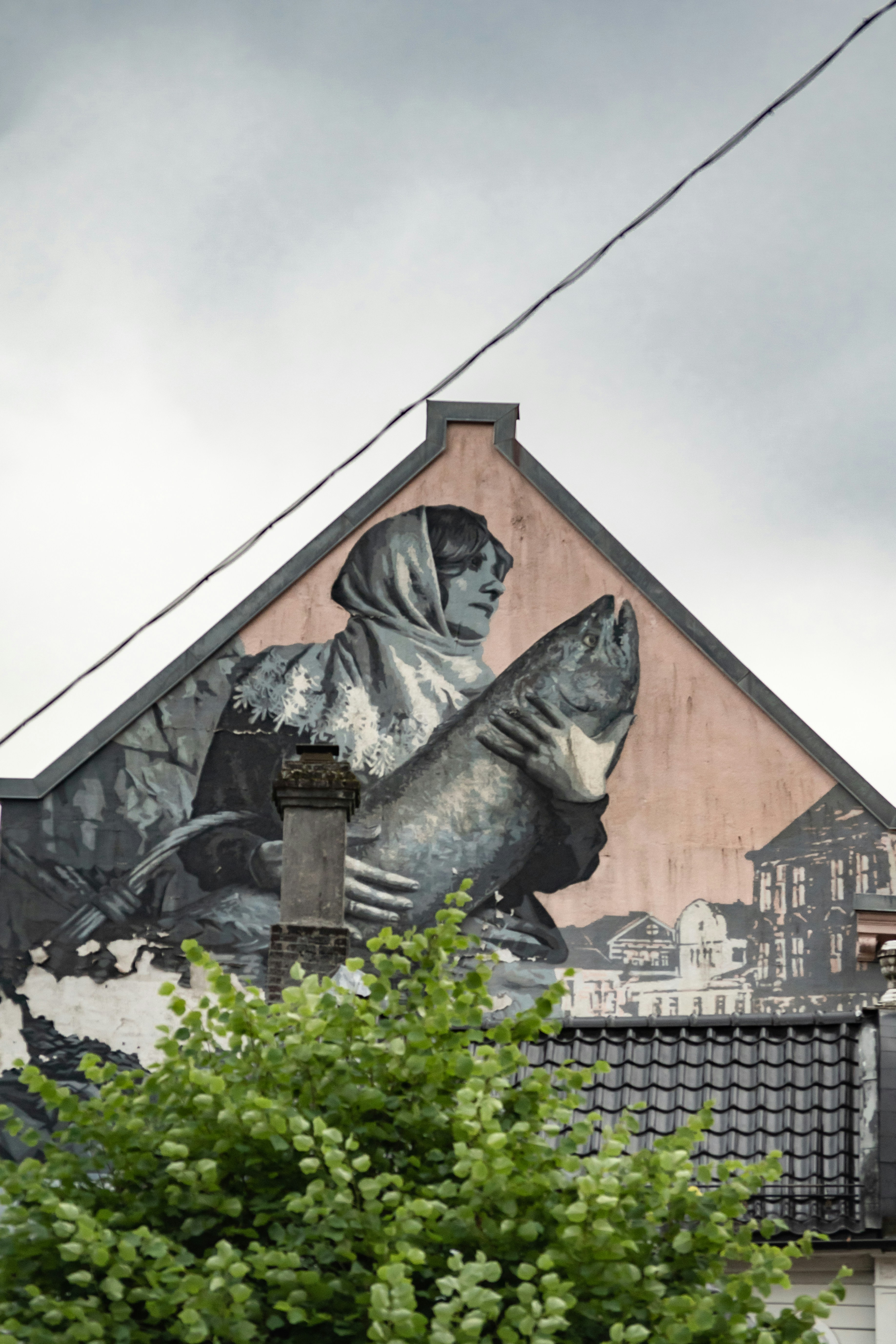 Bildet viser en fasade med et stort veggmaleri av en person som holder en stor fisk. Maleriet er i svart-hvitt, og det er trær og takdetaljer i forgrunnen. «KI-generert bildetekst, kvalitetssikret før publisering.» 