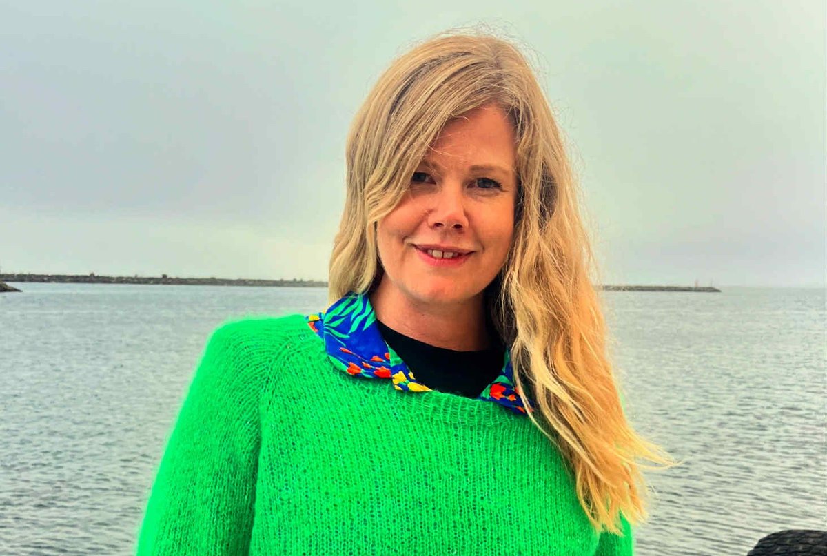 Bildet viser en kvinne med lyst, bølgete hår som smiler mot kameraet. Hun har på seg en grønn genser med en fargerik krage. «KI-generert bildetekst, kvalitetssikret før publisering.» 