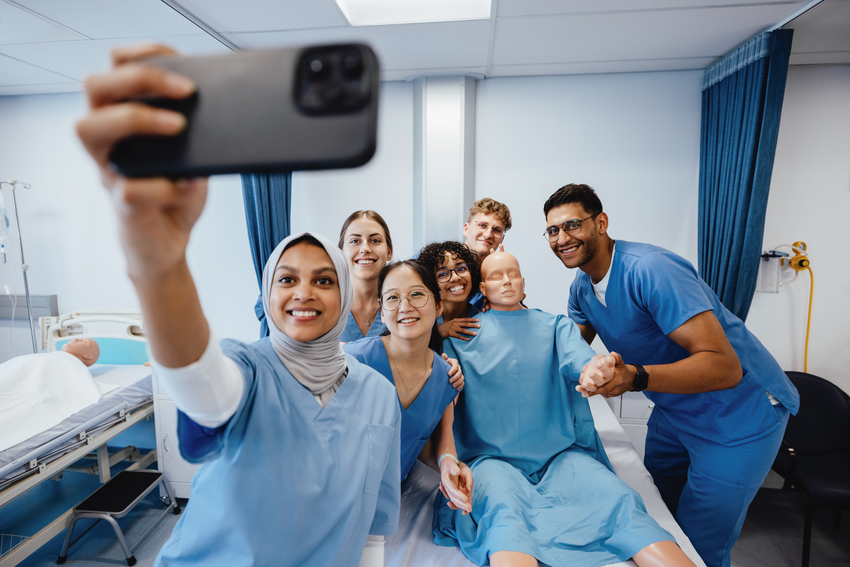 En mangfoldig gruppe av sykepleierstudenter i uniform tar en selfie sammen med en medisinsk treningsdukke i et sykehusrom. De ler og smiler mens de øver seg i praksis, og bildet fanger et øyeblikk av læringsglede, samarbeid og inkludering. «KI-generert bildetekst, kvalitetssikret før publisering.»