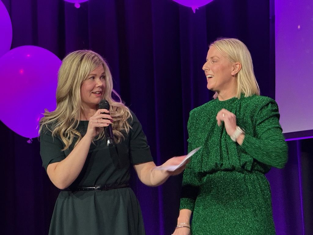 Bildet viser to personer på en scene. Den ene personen holder en mikrofon, mens den andre ser ut til å lytte eller snakke. Begge ser ut til å være glade eller engasjerte. Det er lilla ballonger i bakgrunnen som en del av dekorasjonen. De to er Linn Ceilie Rotvold Bylund og Cathrine Van der Linden fra Steinkjer på scenen under utdelingen av Helseprisen 2025.